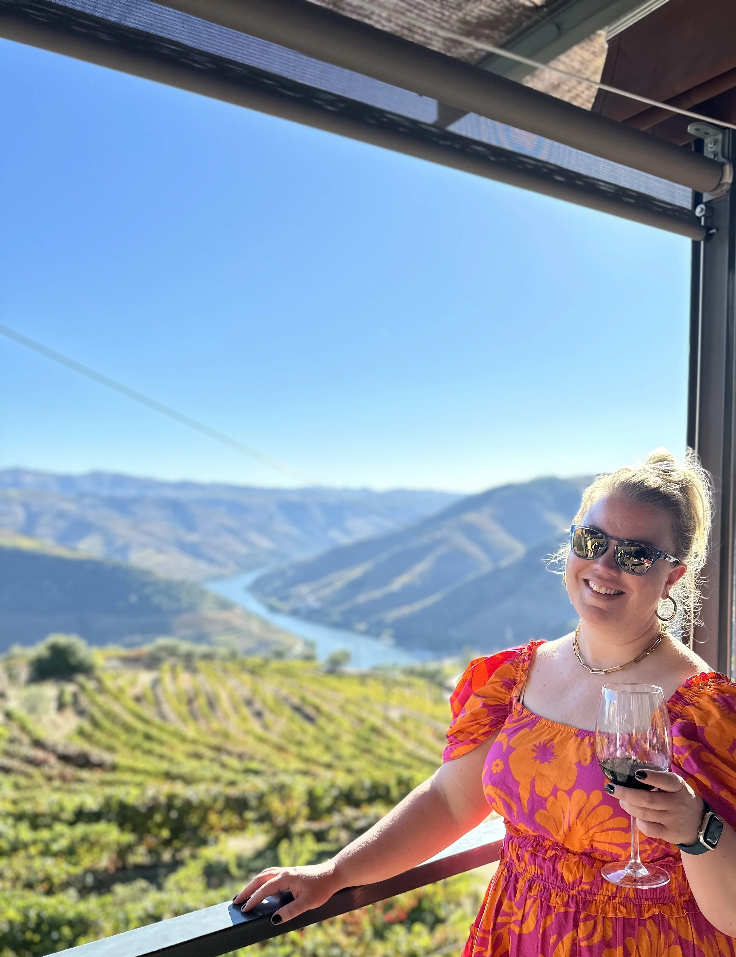 A woman with blonde hair and sunglasses smiling while holding a glass of red wine, standing on a balcony with a scenic view of a river and green hills underneath a clear blue sky in the Duomo Valley outside of Porto.