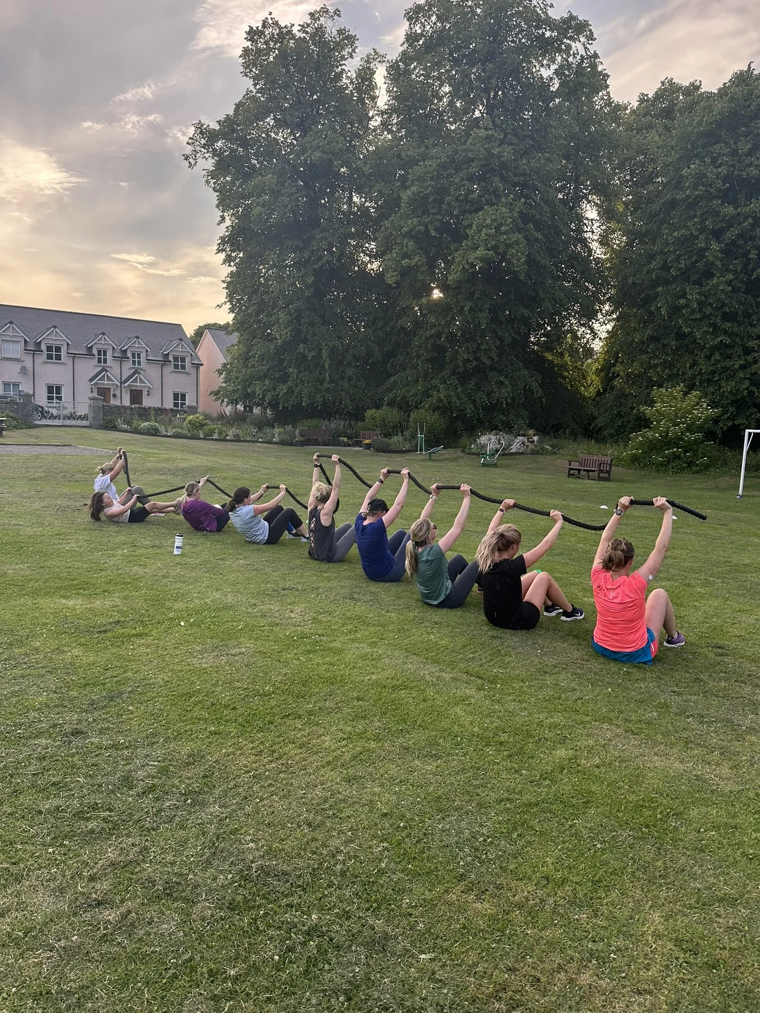 A group of people participating in an outdoor fitness class, sitting on the grass and holding a rope over their heads. The scene is set in a park during sunset with large trees and residential buildings in the background.