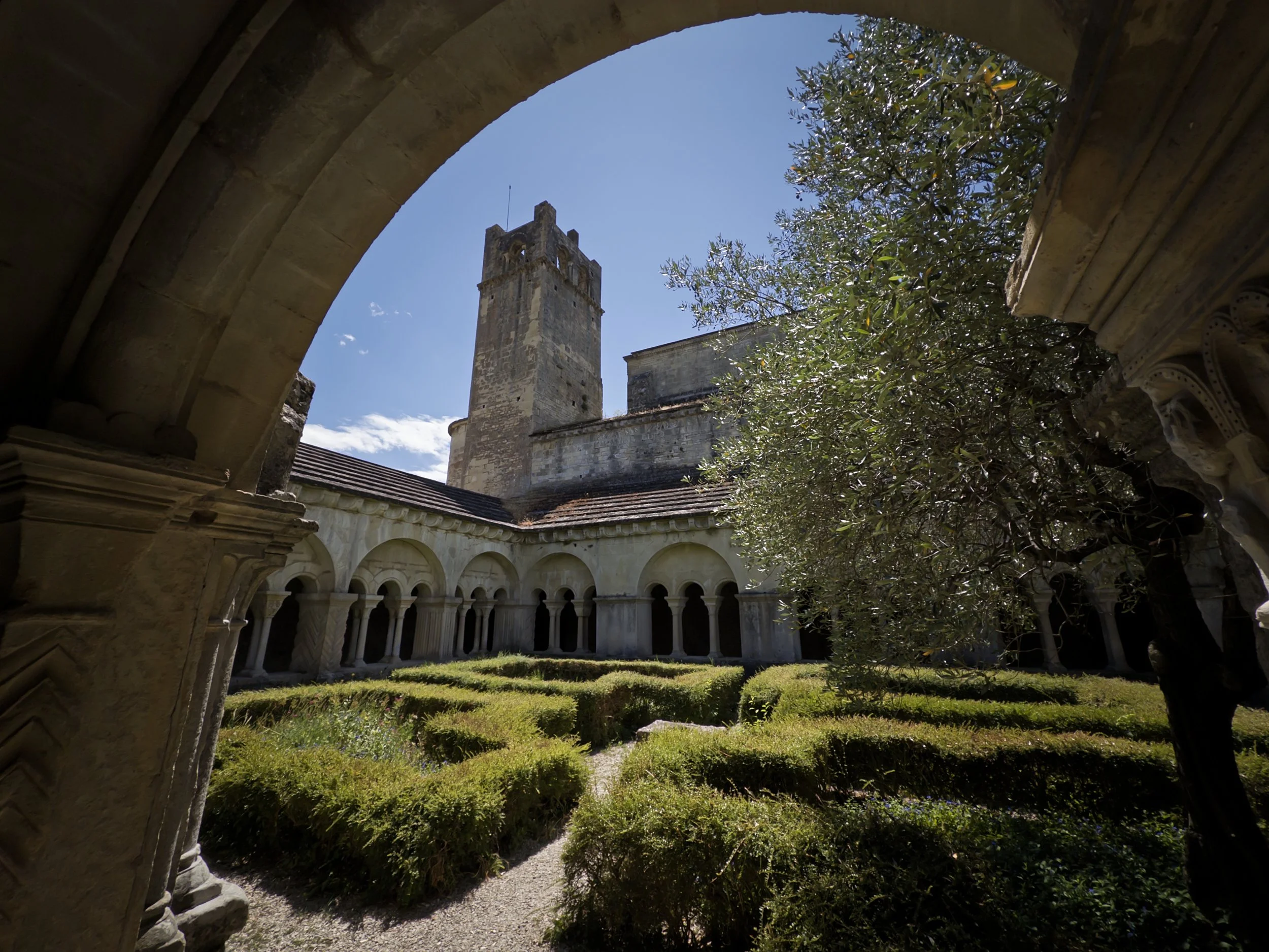 Vue d'un cloître avec de la verdure, des arches en pierre, une tour médiévale en pierre et un ciel bleu avec quelques nuages.