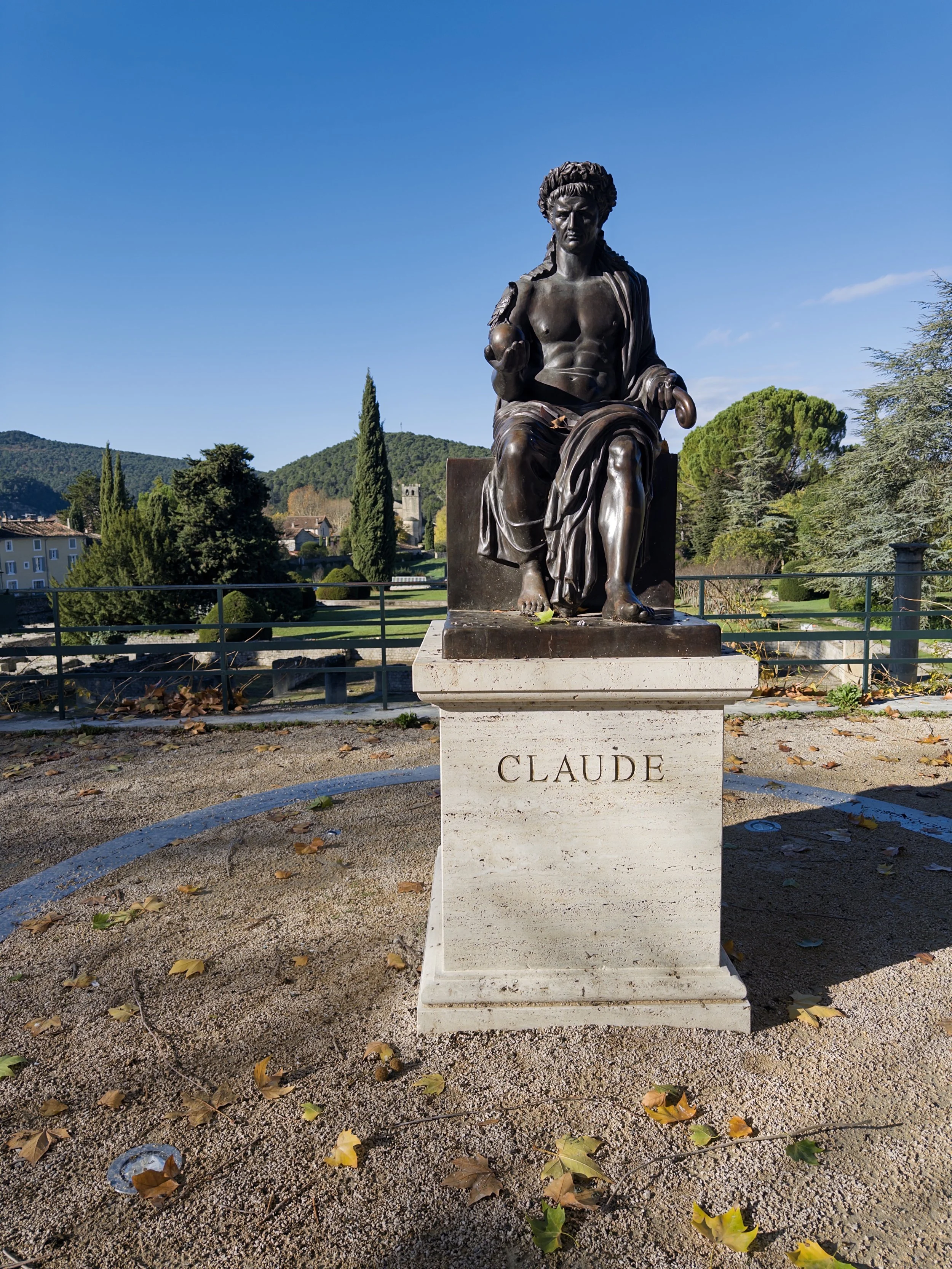 Statue de Claude, représentant un homme assis avec un torse nu et une robe drapée, tenant une boule dans une main et un outil dans l'autre, située sur un socle en pierre dans un parc avec des arbres et une vallée en arrière-plan.
