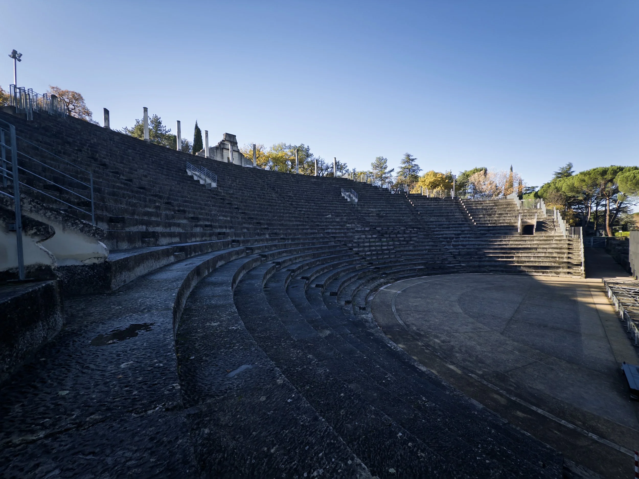 Ancien théâtre romain en pierre avec gradins en demi-cercle, entouré d'arbres et sous un ciel bleu clair.