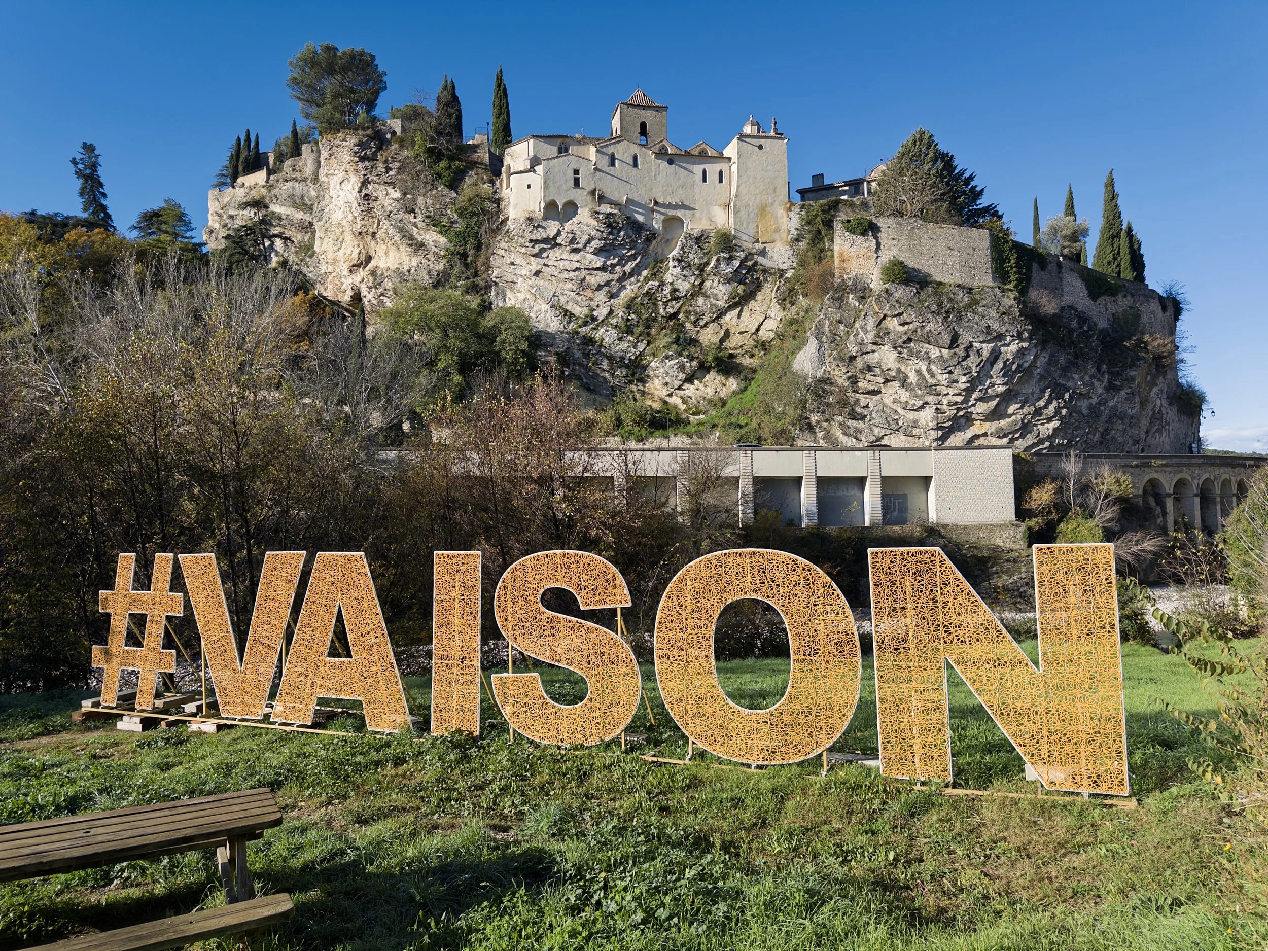 Déclaration d'amour avec un hashtag '#VAISON' en lettres en bois ou en métal dans un jardin avec un château en roche en arrière-plan.