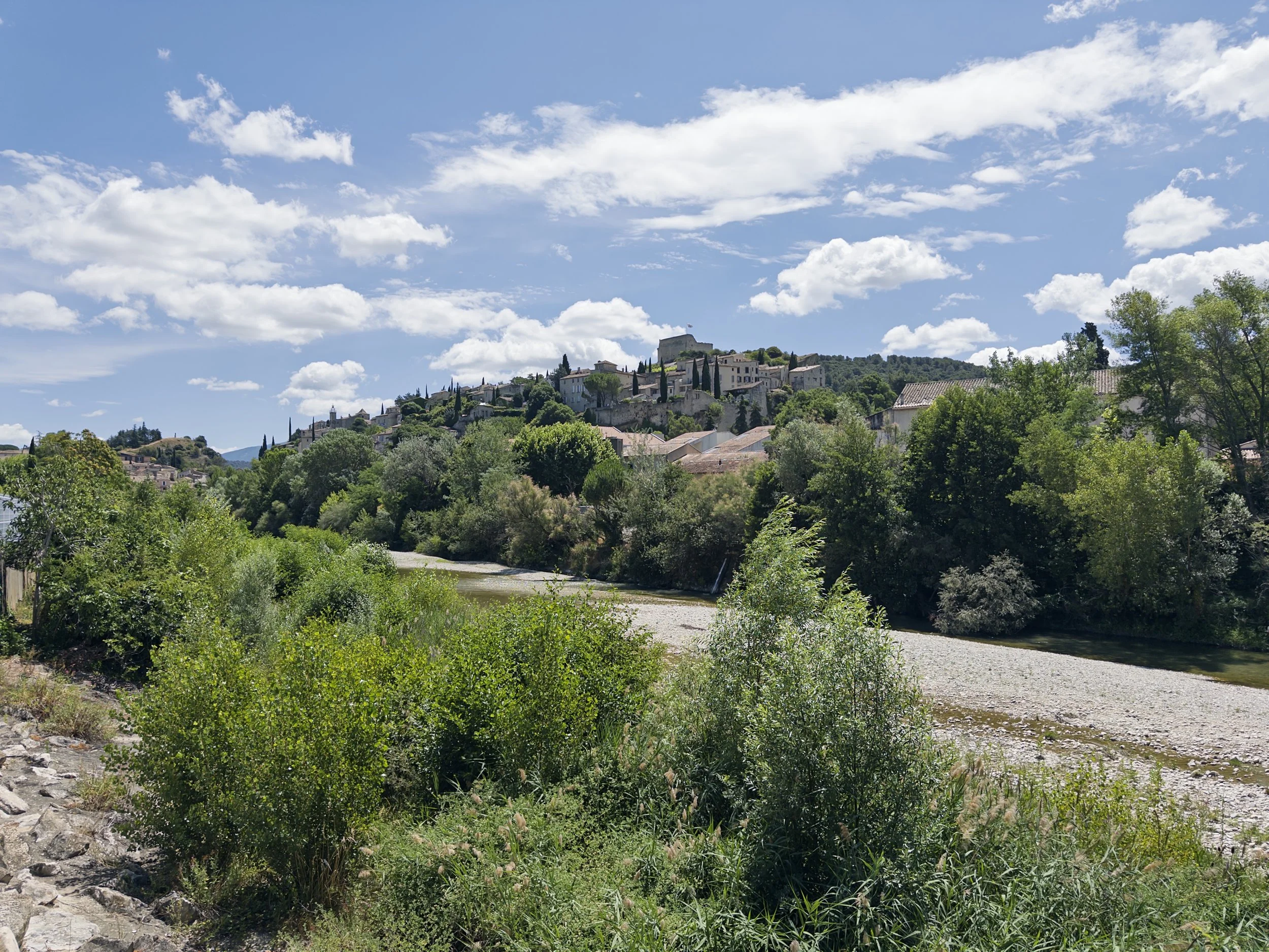 Un paysage avec une rivière bordée d'arbres verdoyants, un village en hauteur avec des maisons en pierre et un ciel bleu avec des nuages blancs.