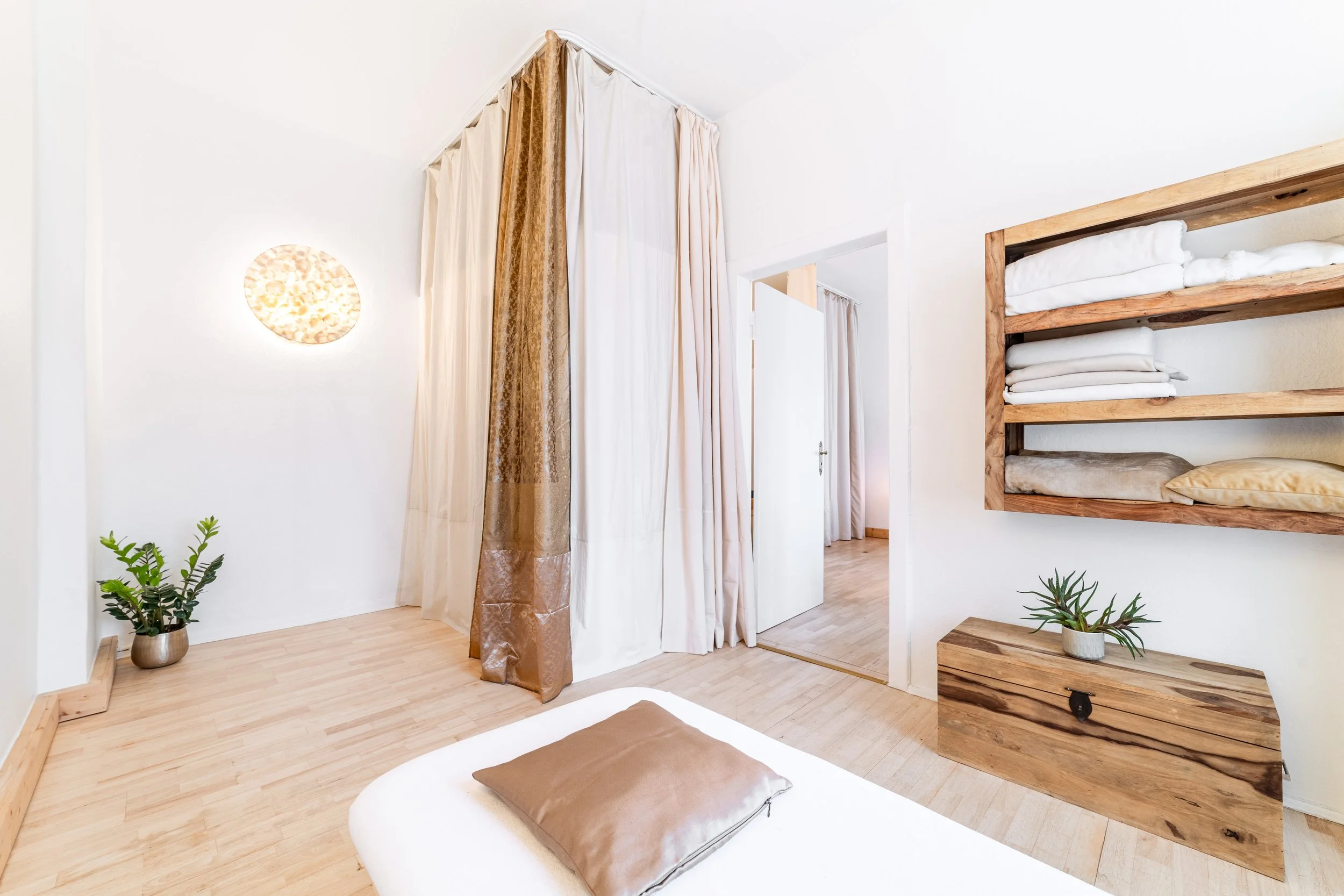 Ein helles Schlafzimmer mit Holzboden, Bett mit brauner Kissen, einen kleinen Holzkasten mit Pflanze, Wandregal mit weißen und gelben Betten und einem Vorhang im Raum.