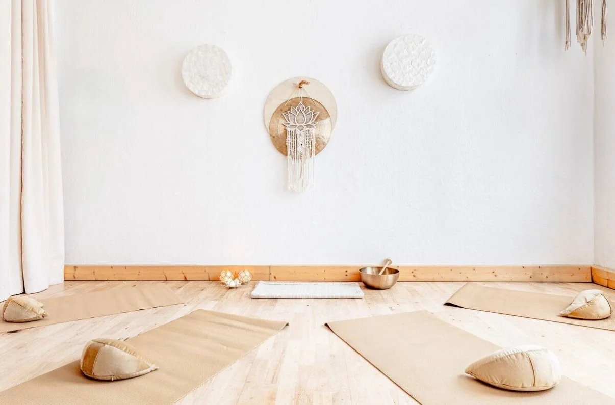 Ein Raum für Meditation mit Matten, Kissen, einer Klangschale und Dekorationen an der Wand in neutralen Farben.
