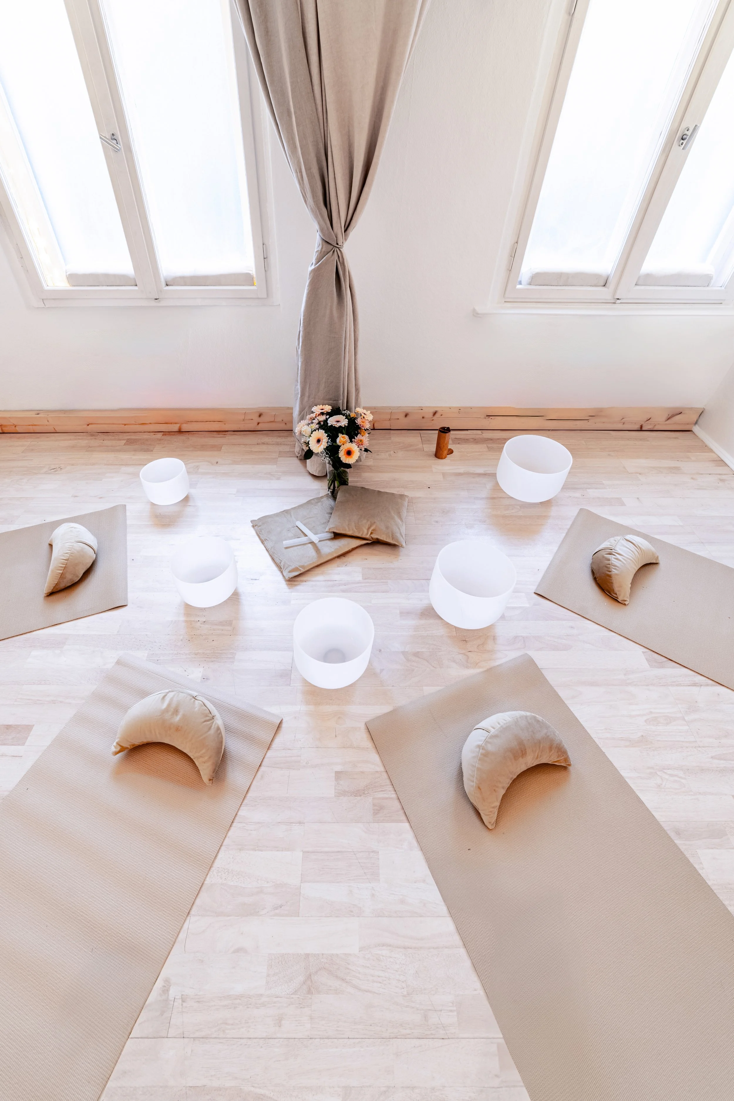 Ein Yoga- oder Meditationsraum mit fünf Yogamatten, Kissen, Kerzen, einer Vase mit Blumen und einem Klangschale vor großen Fenstern mit beige Vorhängen.