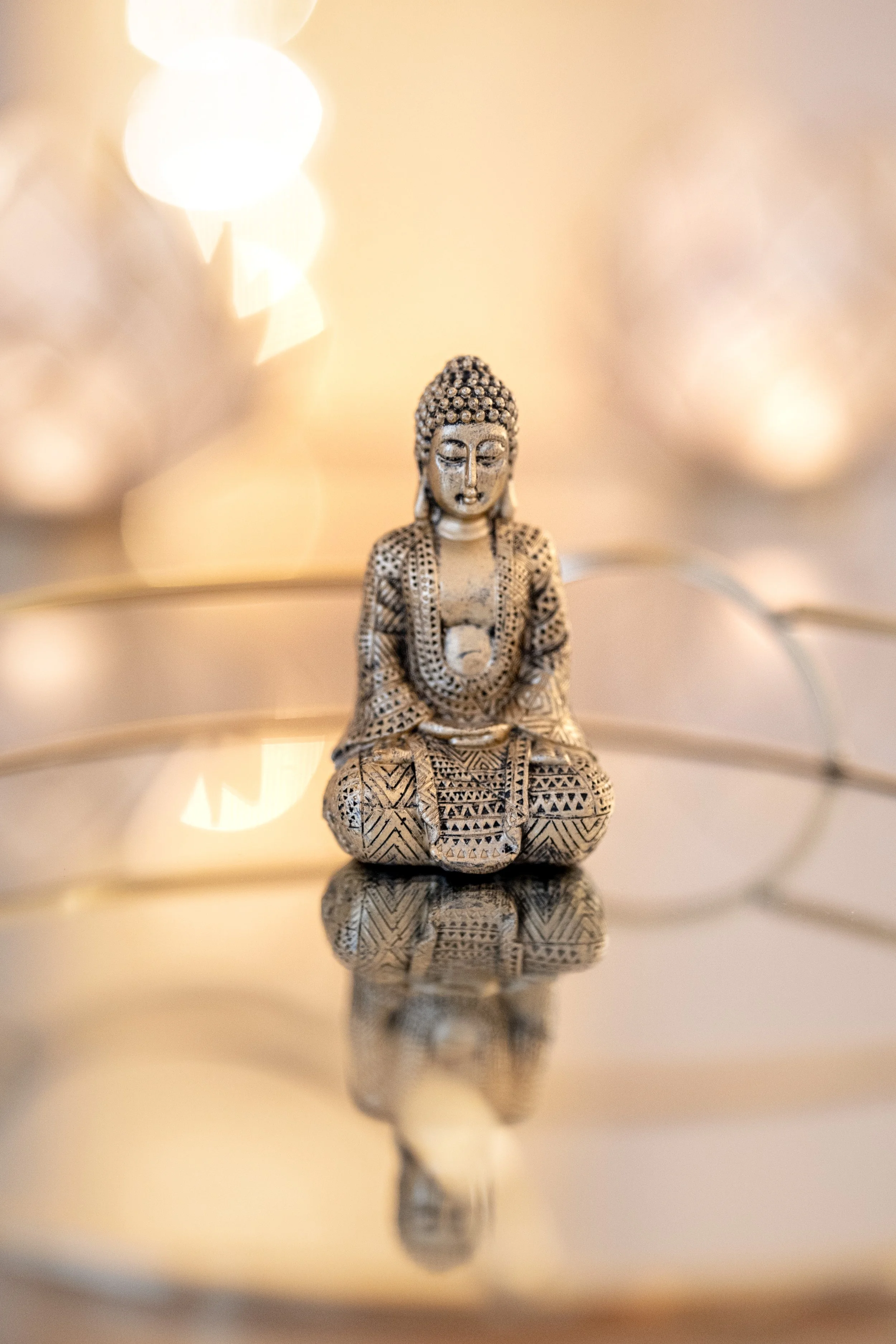 Kleine Buddha-Figur aus silberfarbenem Material mit detaillierten Mustern, sitzend auf spiegelndem Untergrund, mit warmem, verfälschtem Hintergrundlicht.