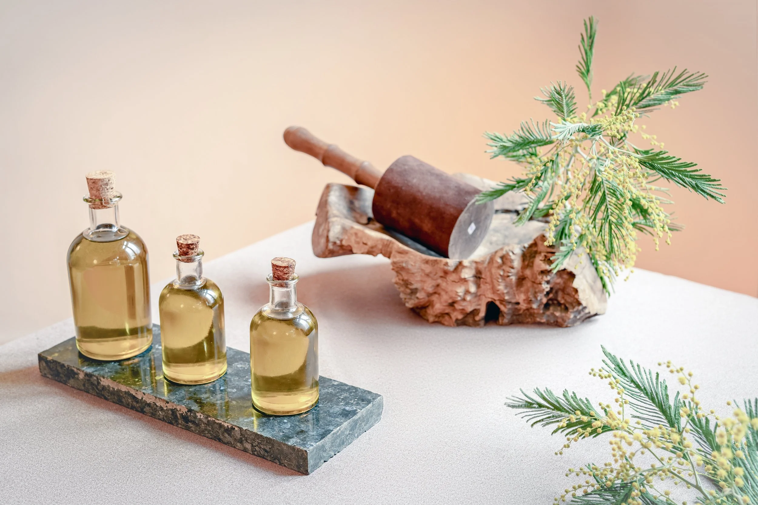 Drei Fläschchen mit goldener Flüssigkeit auf einem schwarzen Marmorblock, daneben Holzscheite, ein Mörser mit Stößel und grüne Zweige mit gelben Blüten.
