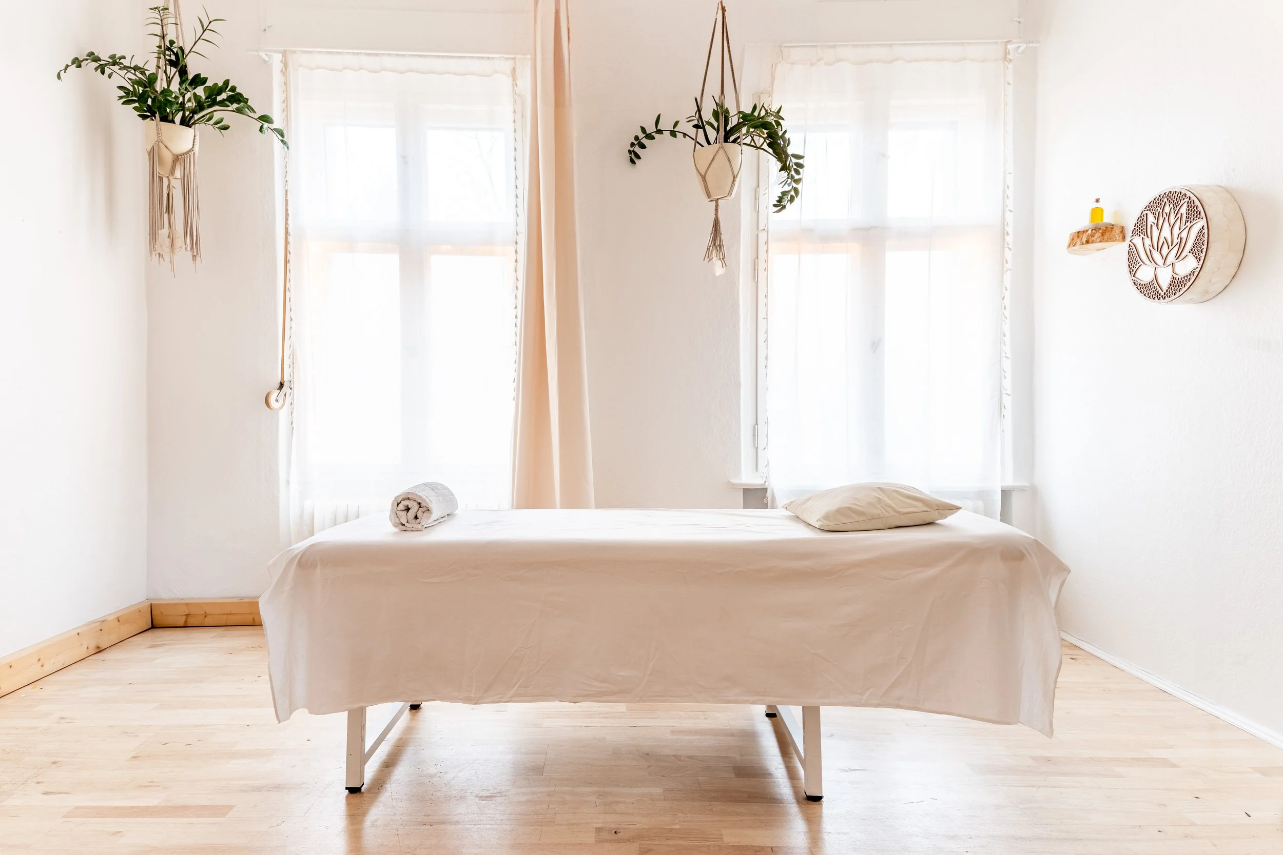 Ein weißes Zimmer mit einem Massagetisch, weißem Bettwäsche, Kissen und Handtuch, zwei Fenster, hängende Pflanzen und Wanddekoration mit Lotus-Design.