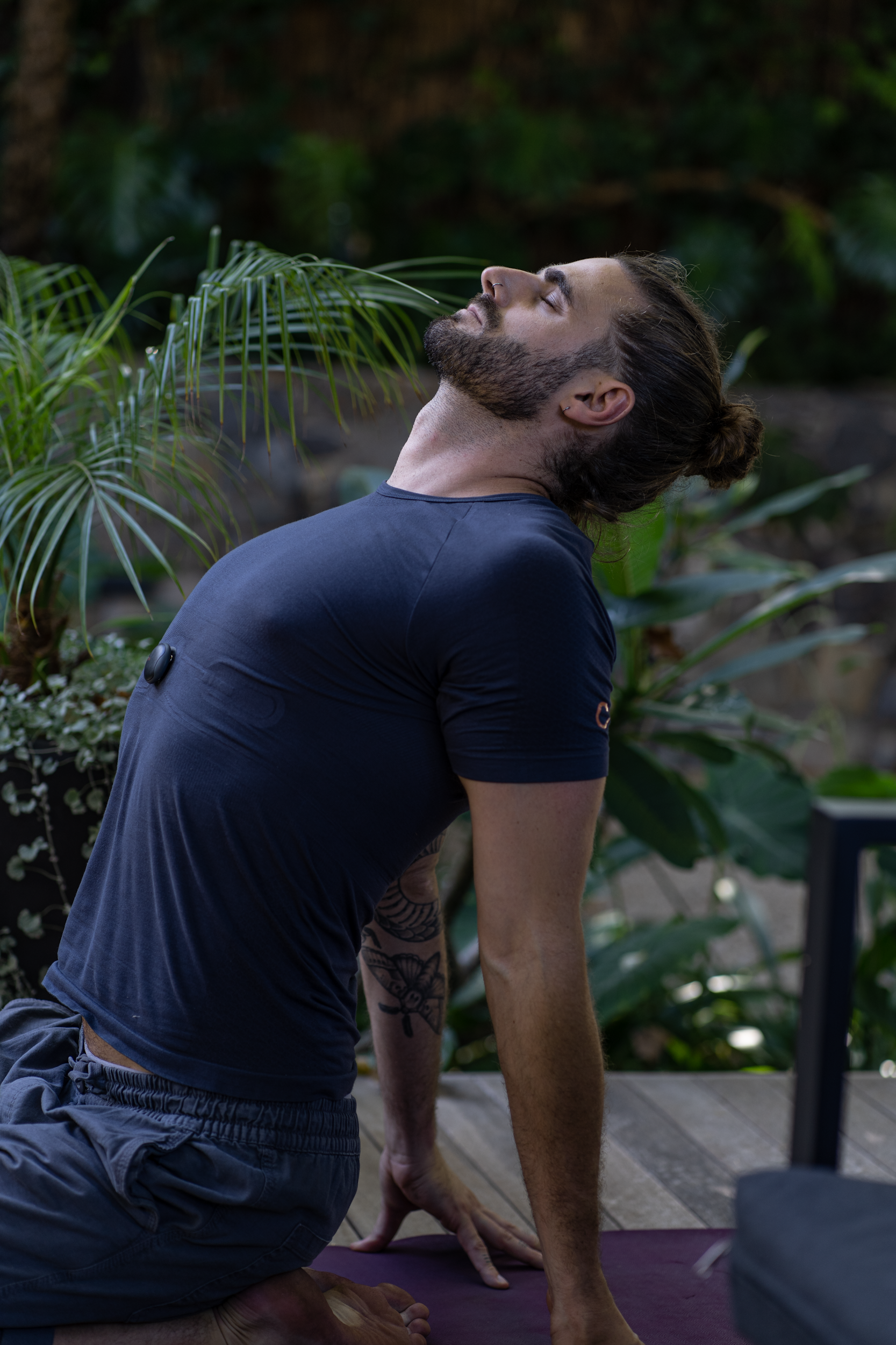 Man with a beard and tattoo on arm practicing yoga outdoors on a purple mat, with tropical plants in the background.