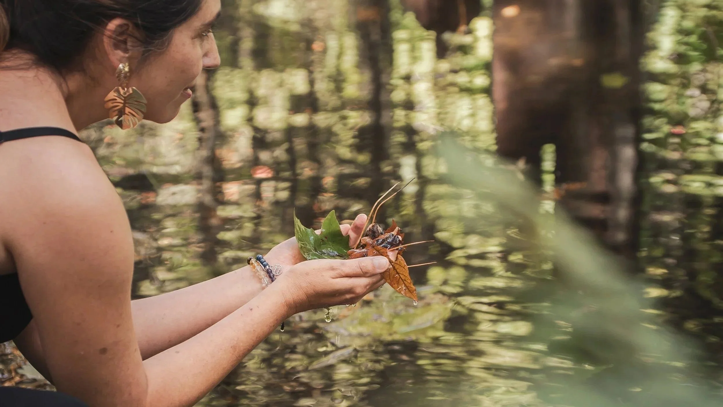 Une femme tenant des feuilles et des éléments naturels près de l'eau dans une forêt.