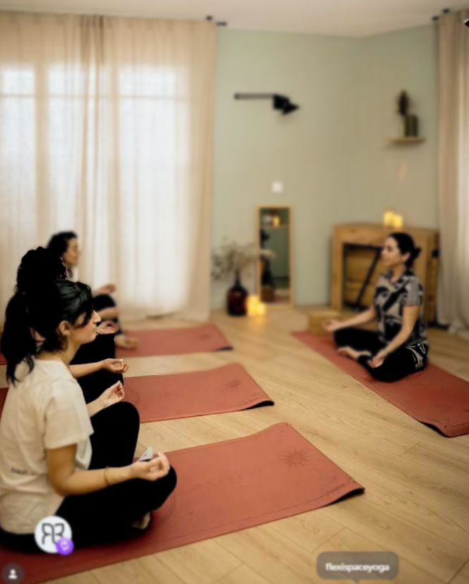 Cinq personnes pratiquant le yoga ou la méditation dans une salle calme avec des tapis de yoga. Une femme mène la séance, assise en tailleur devant un mur décoré.