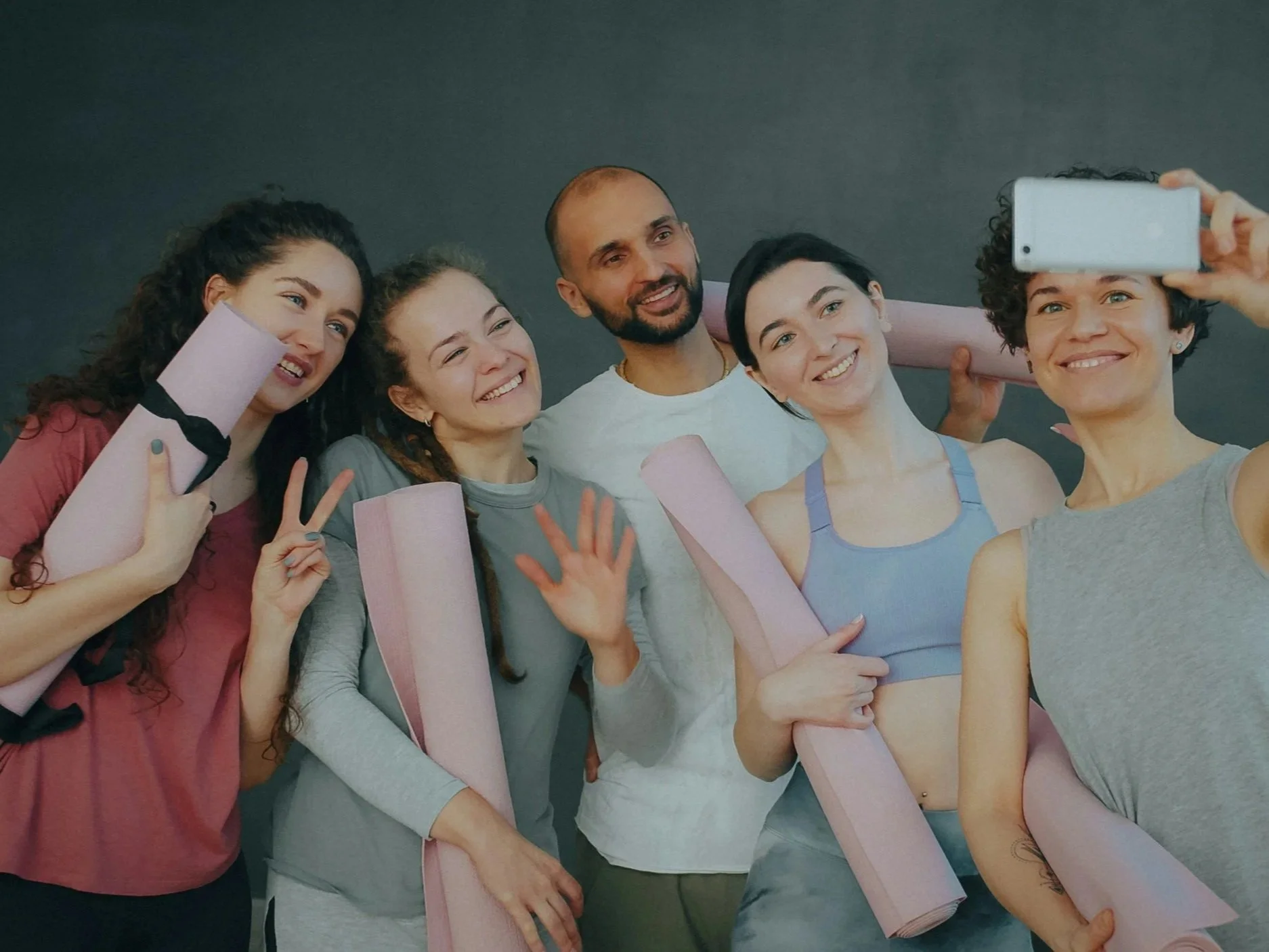 Un groupe de six personnes souriantes posant pour une photo, certains tenant des rouleaux de yoga et faisant des gestes de paix, tandis qu'une personne à droite prend une selfie avec son téléphone.