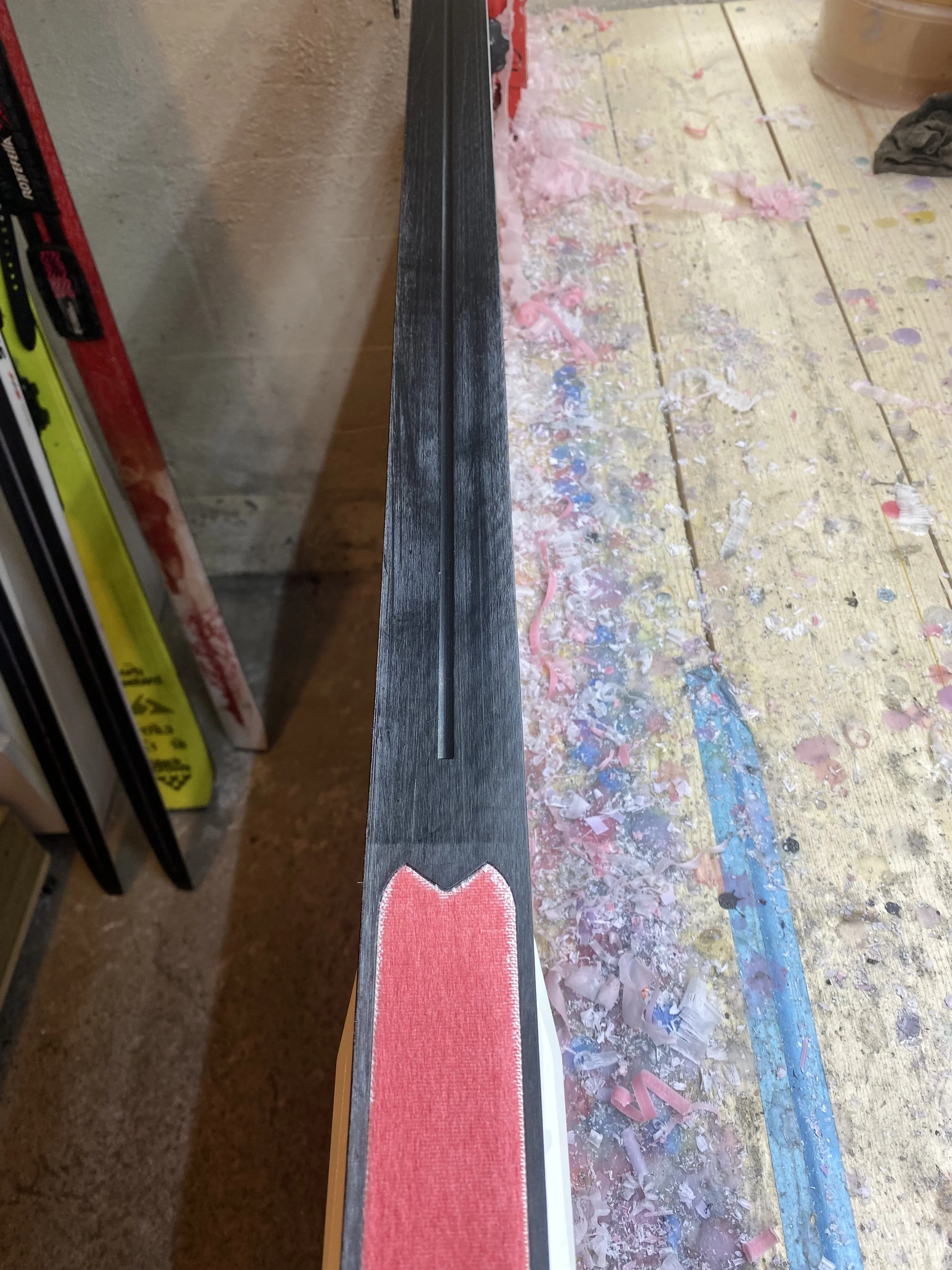 Nærbilde av en lang, svart ski med en rødt merket spore og omtrentingsvis en liten rille i midten. Ski ligger på en verkstedbenk med mye fargede sagflis og plastavfall rundt.