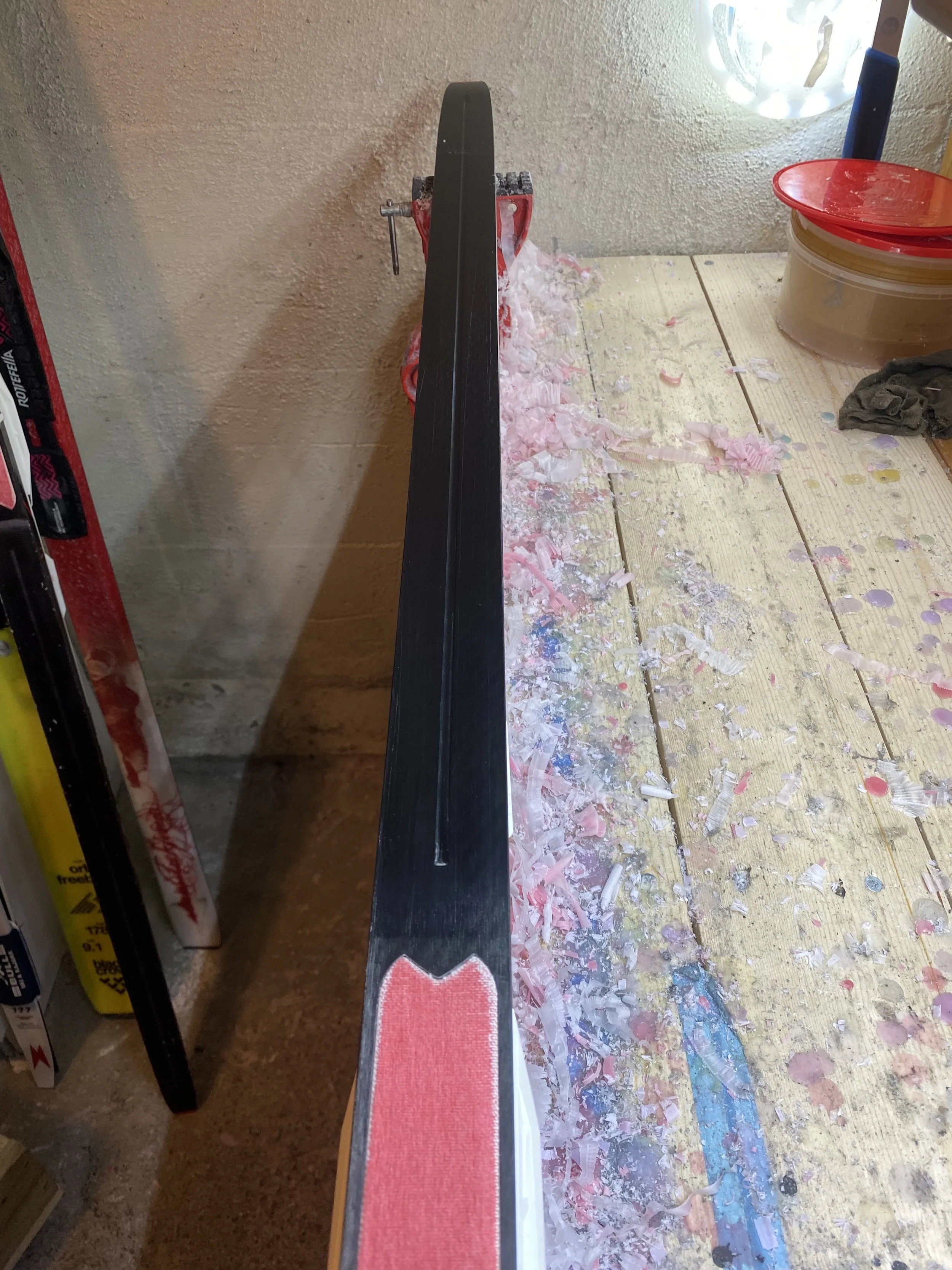 Skihockeykølle med sort skaft og rødt grep, plassert på et arbeidsbord med papp- og skøytebellallekningsrester.