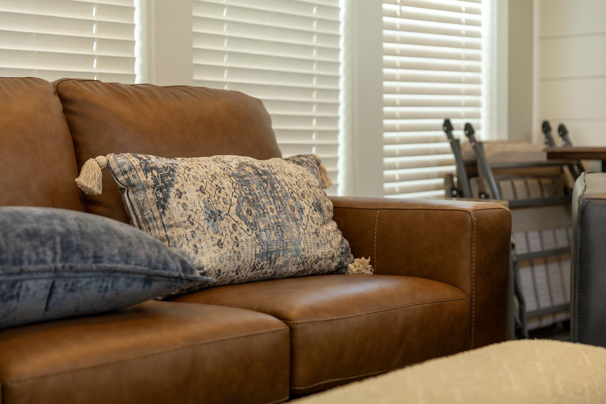 Close-up of a brown leather sofa with patterned pillows in a room with white blinds.