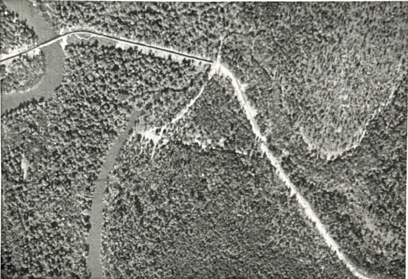 Satellite image showing a winding road passing through a heavily forested area with clearings and small bodies of water nearby.