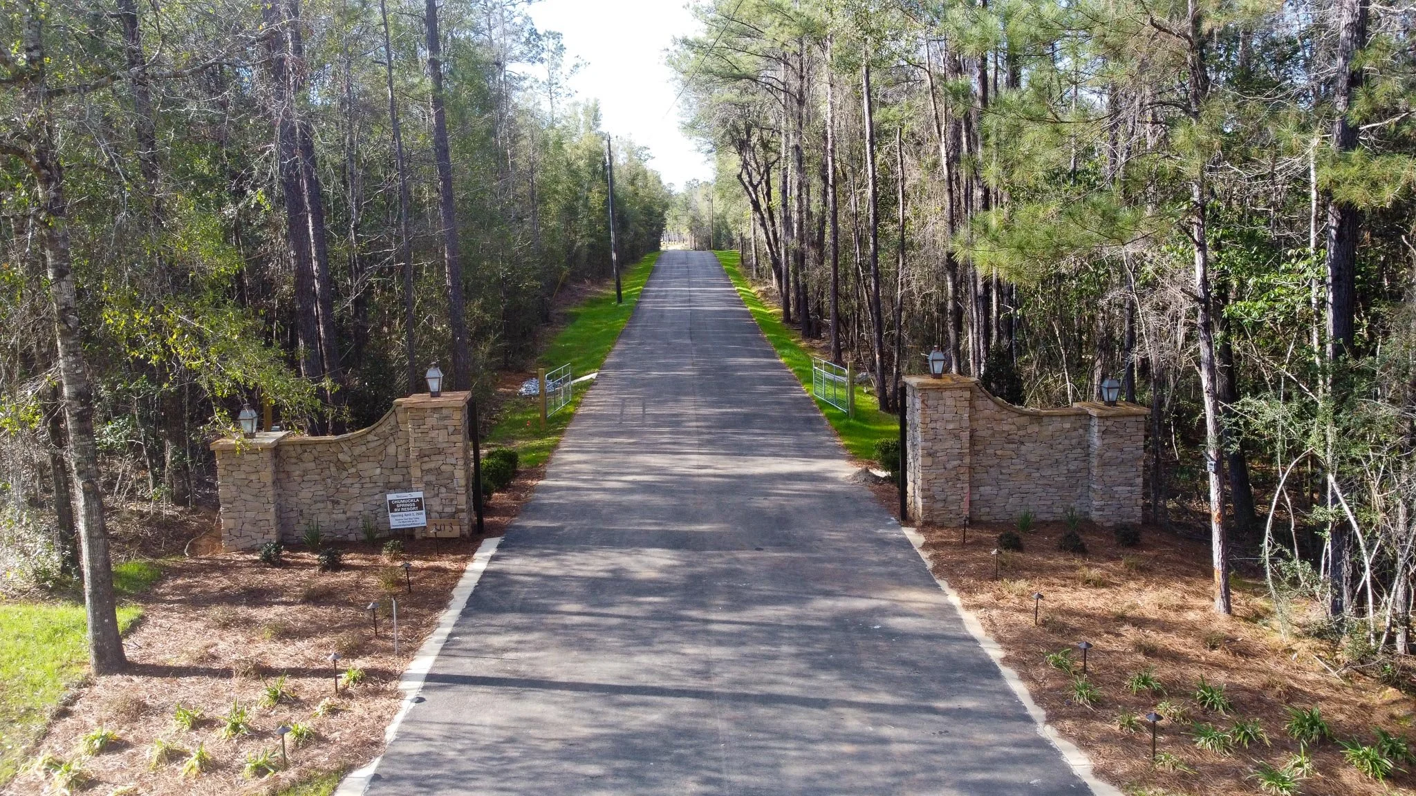 Aerial view of a park with winding roads, green lawns, trees, small buildings, and surrounding water.