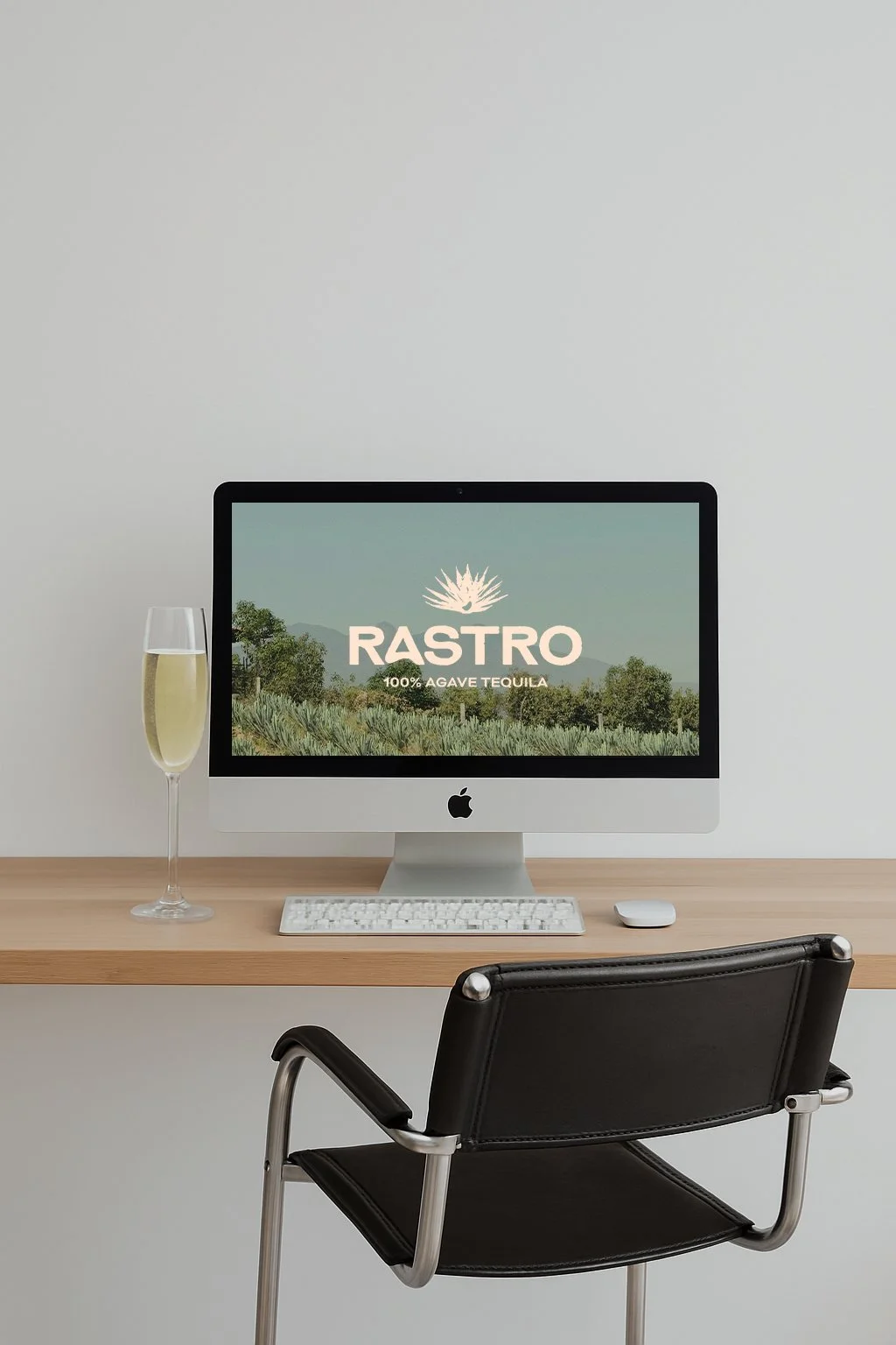 An Apple iMac on a wooden desk displaying a website for Rastro, a 100% agave tequila, with a glass of champagne beside it and a black office chair in front.