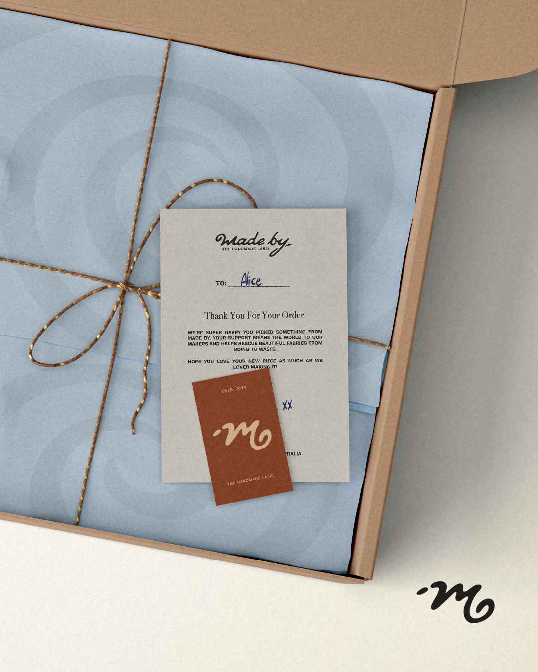 Open gift box with a blue tissue paper, a handwritten note, and a small brown tag with a white logo, tied with a gold and copper string.