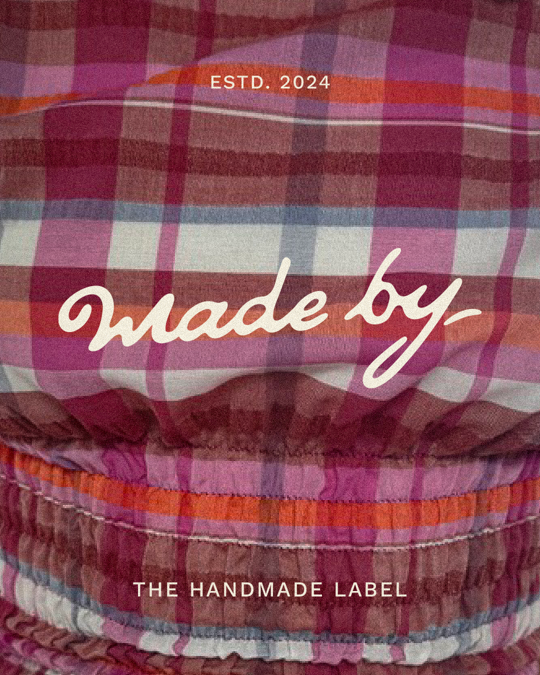 Close-up of fabric with pink, purple, and orange plaid pattern, featuring white text that reads 'made by' and 'the handmade label' with a small text at the top that says 'estd. 2024'.