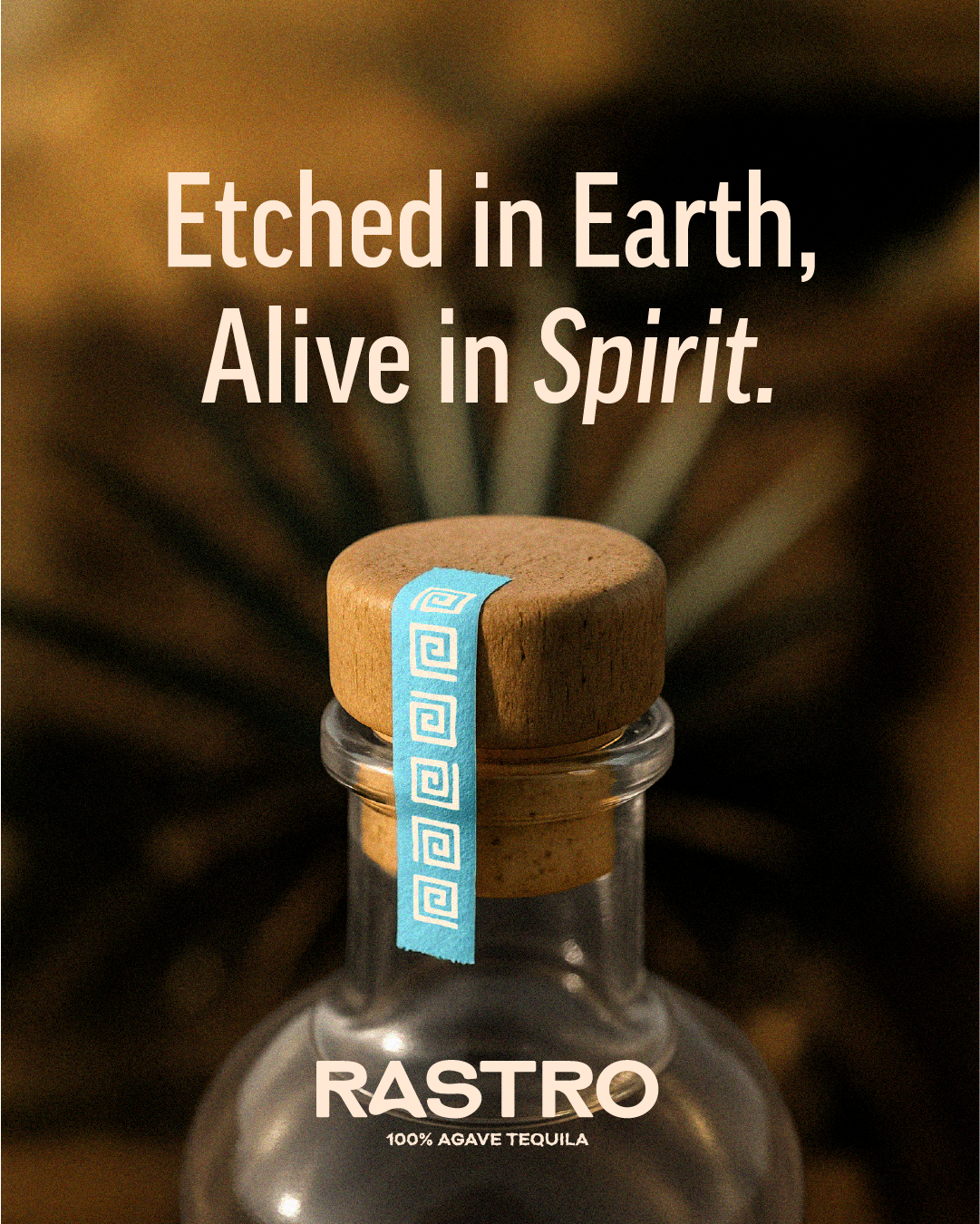 Close-up of a glass bottle with a wooden stopper, sealed with a light blue tape featuring white spirals, against a blurred, earthy background. Foreground text reads, 'Etched in Earth, Alive in Spirit.' and the bottom text says, 'RASTRO 100% AGAVE TEQ