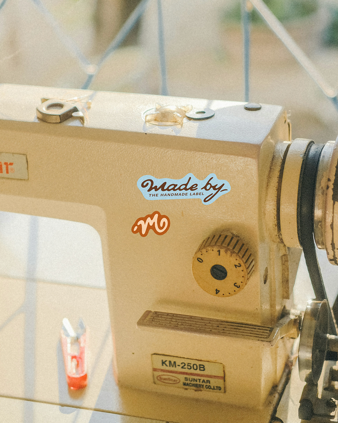 Close-up of a beige sewing machine with stickers that say "Made by the handmade label" and a stylized "M" logo. Part of the machine's dial and a small container on top are visible.