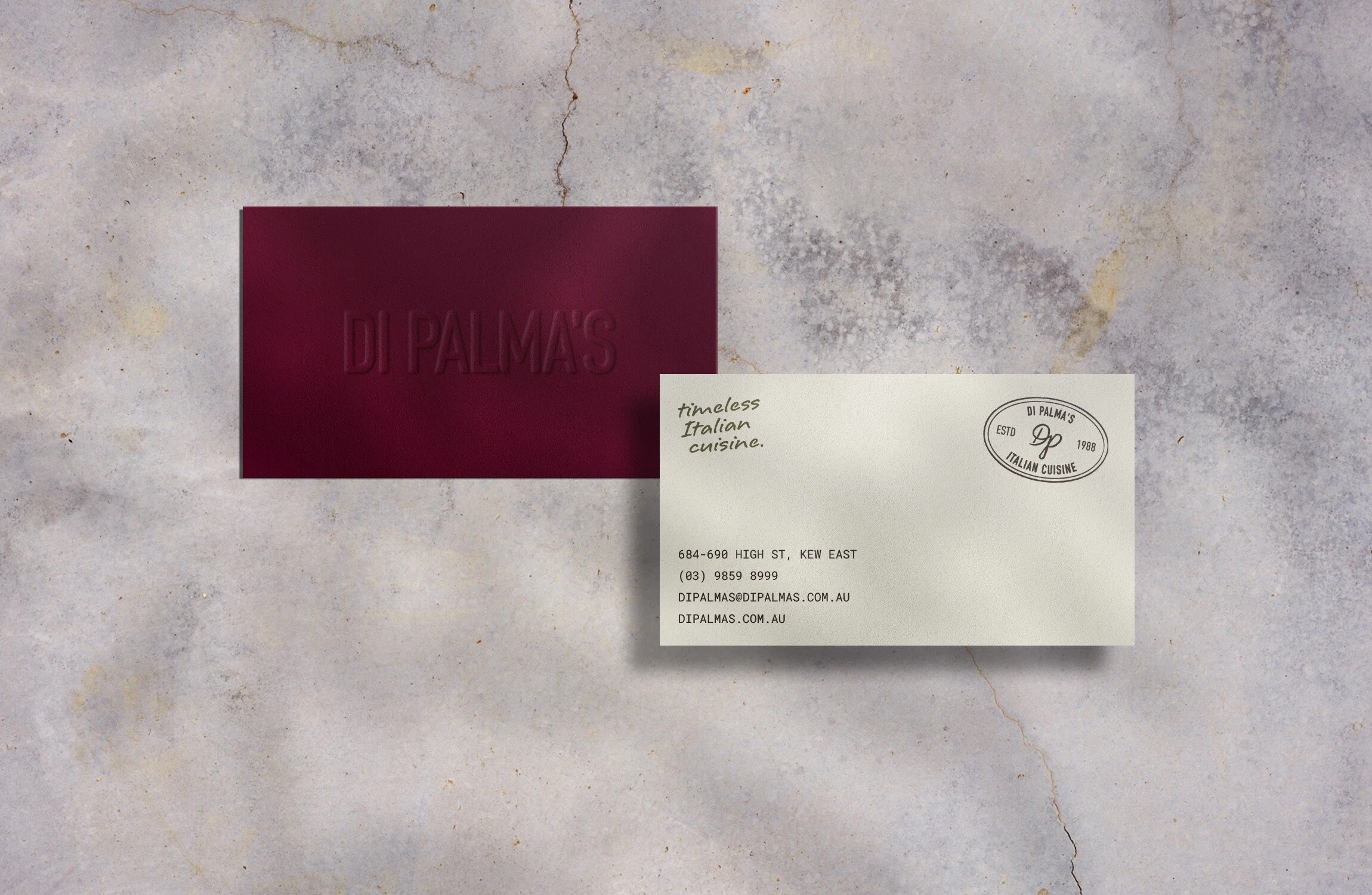 A burgundy business card with embossed text 'DI PALMA'S', a cream-colored card with handwritten style text 'timeless Italian cuisine', and a round logo with 'DI PALMA'S', 'ESTD', '1988', and 'ITALIAN CUISINE', all placed on a light grey, textured concrete surface.