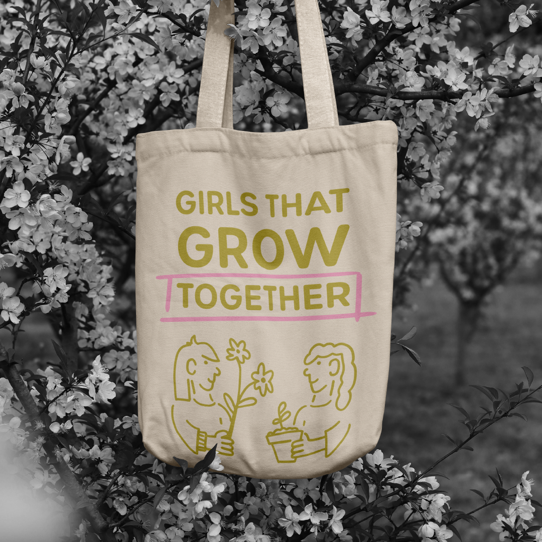 A beige tote bag hanging on a branch of a flowering tree with white blossoms. The bag has text that reads "Girls That Grow Together" with the word "Grow" in large font and a pink underline under the word "Together." There are simple line drawings of