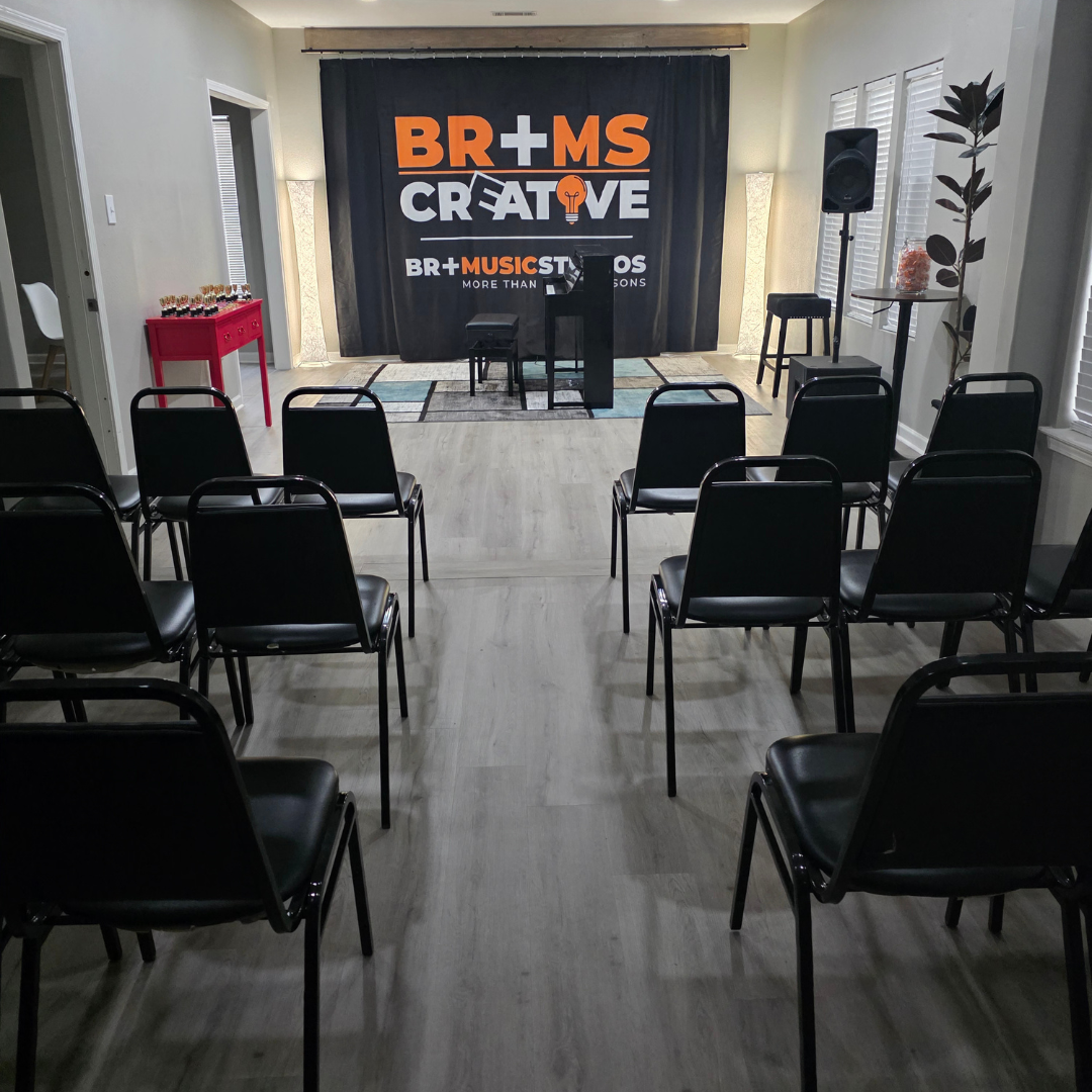 Empty indoor stage area with chairs arranged in rows facing a black backdrop with the text "BR+MS CREATIVE" and a lightbulb icon, indicating a performance or presentation space.