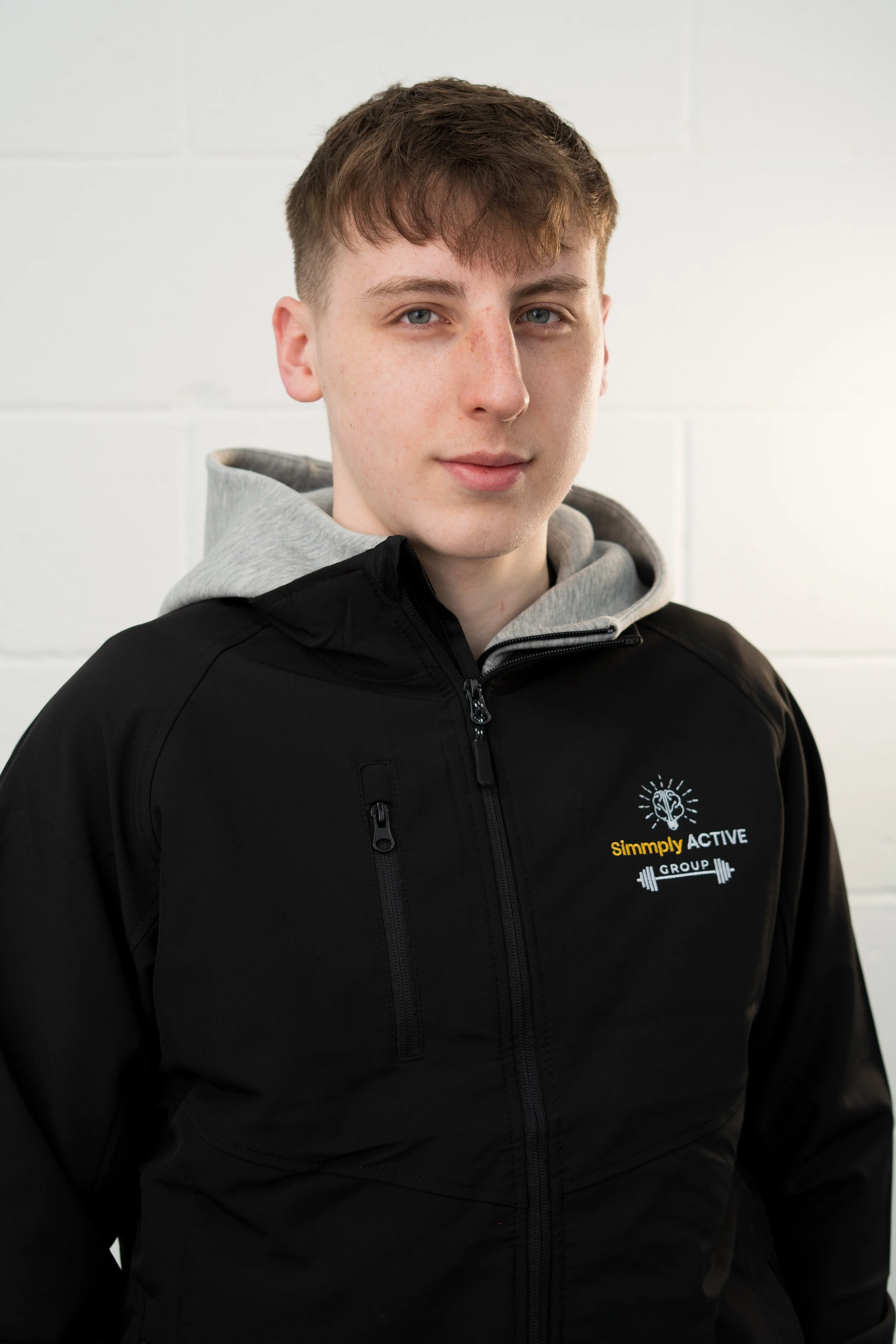 A young man with short brown hair and a light mustache, wearing a black zip-up vest with the 'Simply Active' logo, standing in front of a white backdrop with the same logo and institutional illustrations.