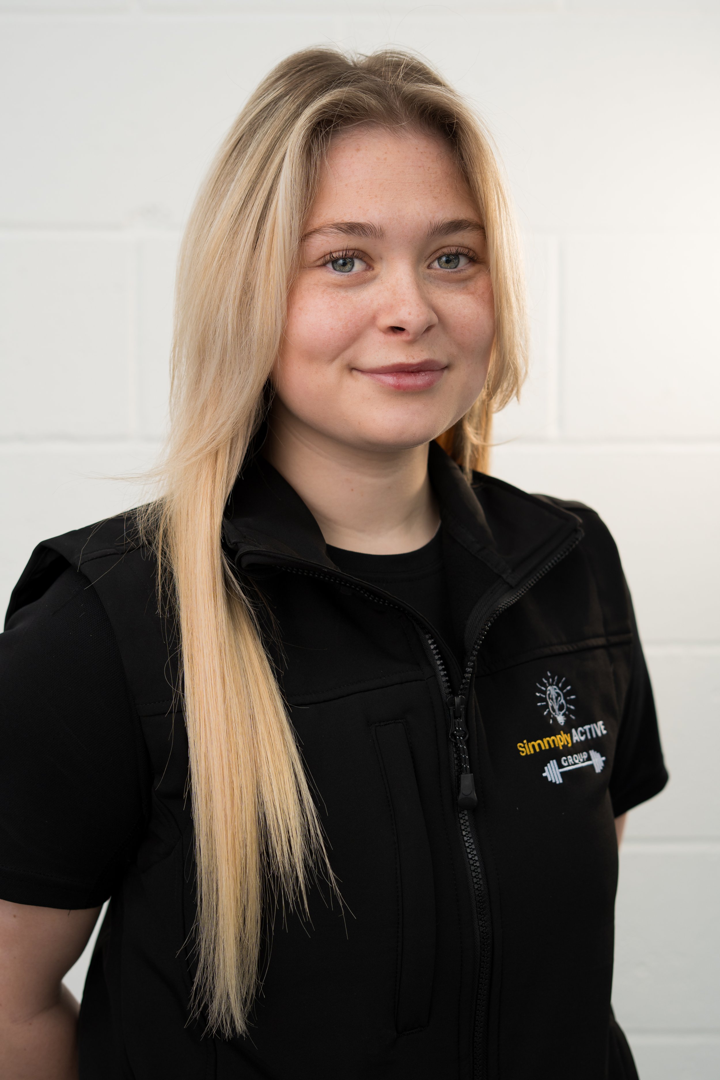 A woman with long blonde hair wearing a black T-shirt with the logo for Simply Active Group, standing in front of a white background with repeating logos of a brain and dumbbell, and the words Simply Active.
