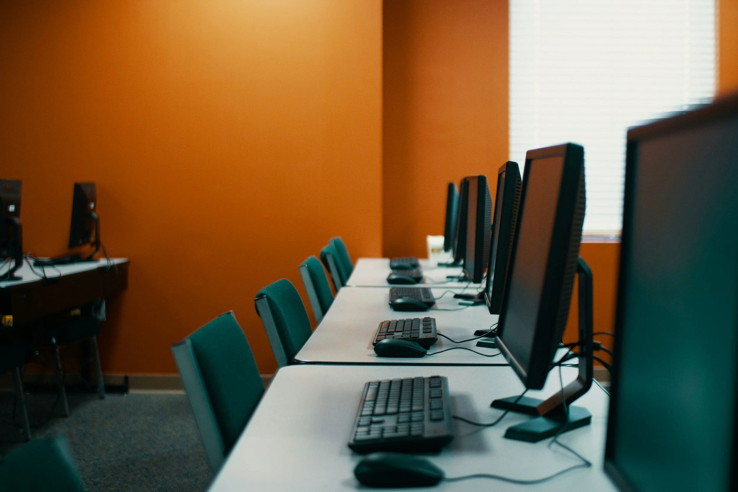 A row of computers with monitors, keyboards, and mice on white desks in a computer lab or classroom, with green chairs and an orange wall in the background.