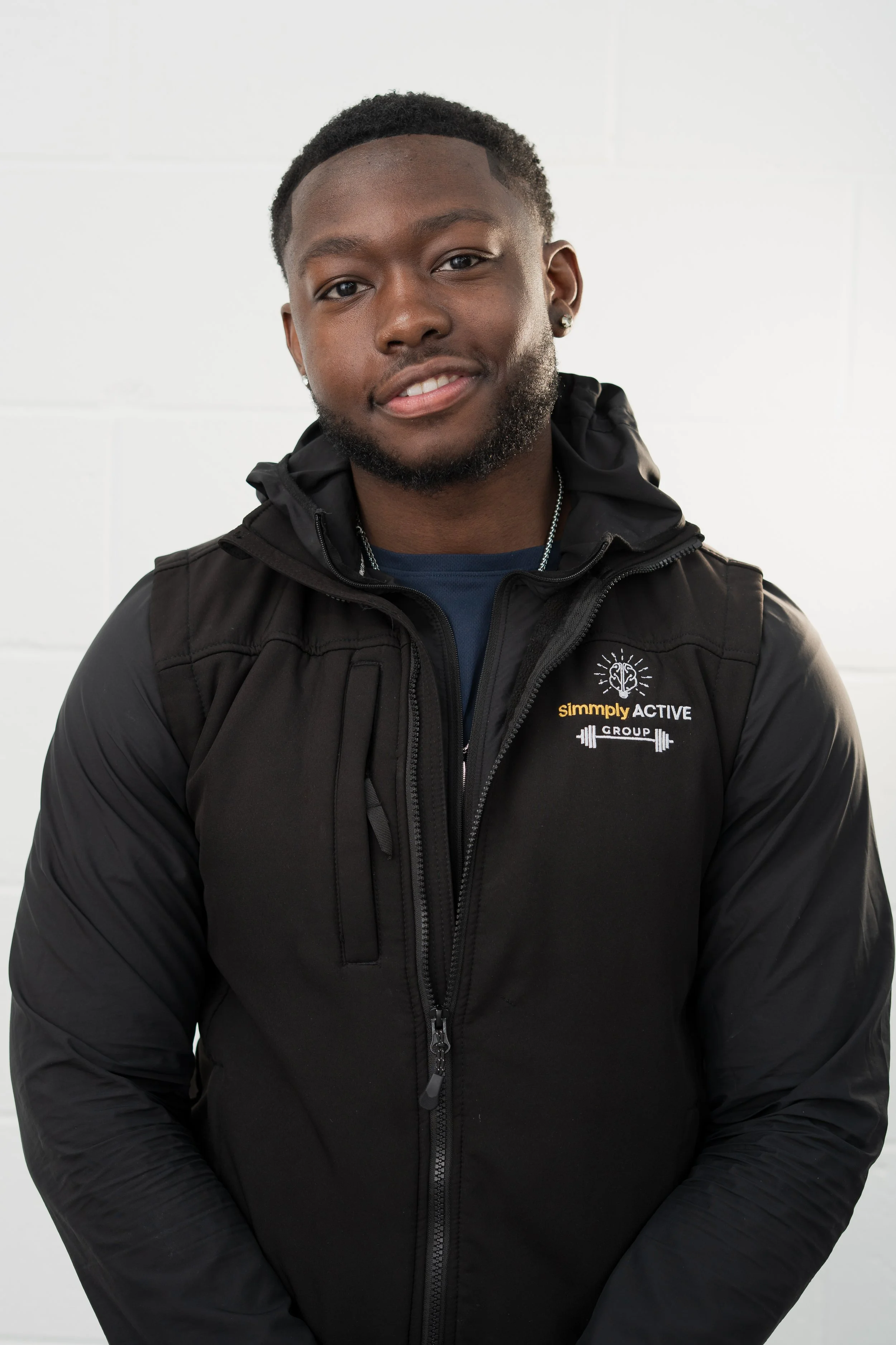 A young man with glasses and a beard, standing in front of a white background with the 'Simmly ACTIVE' logo and graphics. He is wearing a black vest with the logo, a black T-shirt, and black pants.