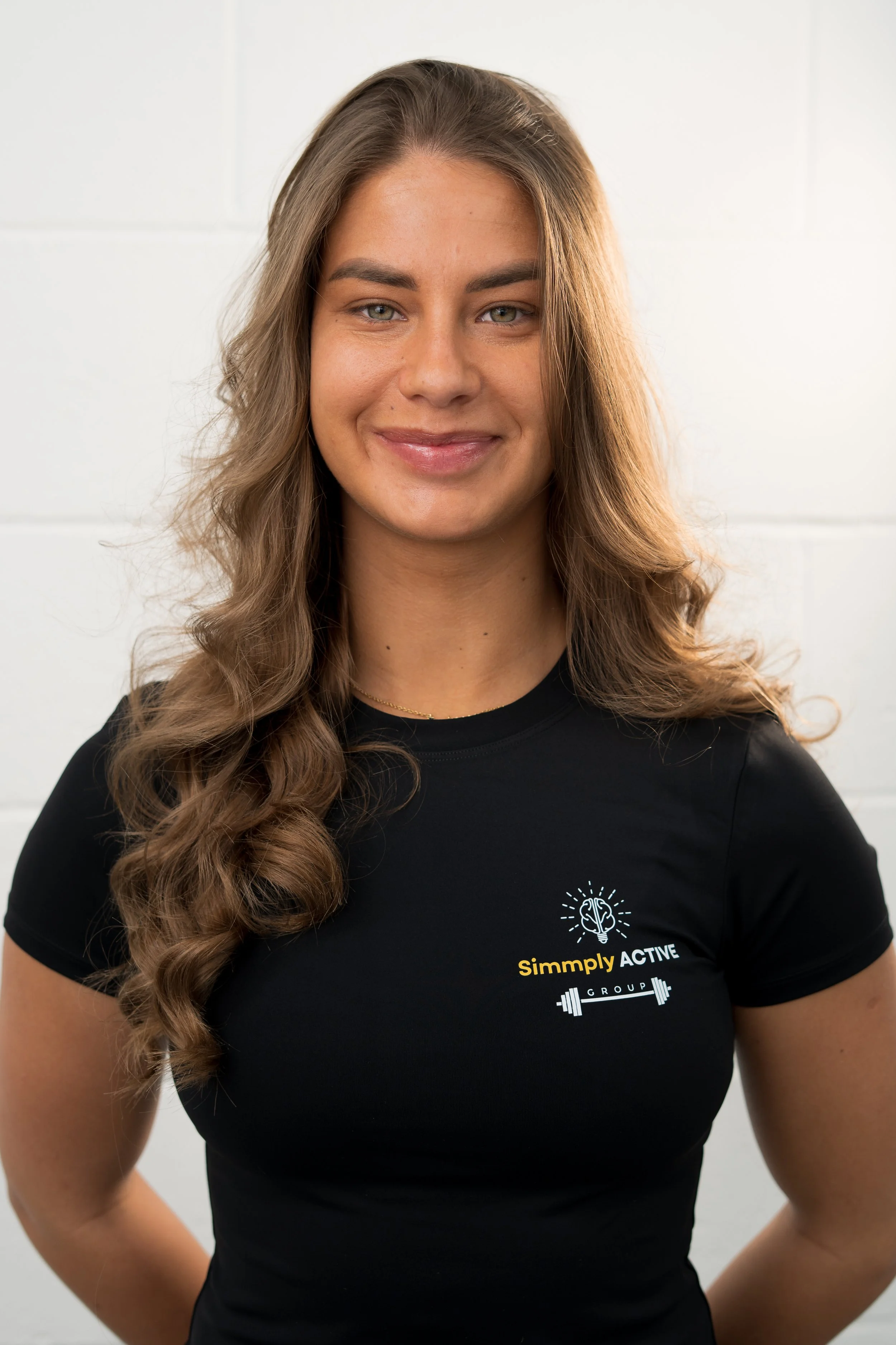 A young woman with long wavy brown hair and light skin, smiling, stands in front of a white background with logos and text that read 'Simply ACTIVE GROUP' and feature a brain illustration and a barbell. She is wearing a black T-shirt with a similar logo on the left side of her chest and black pants.