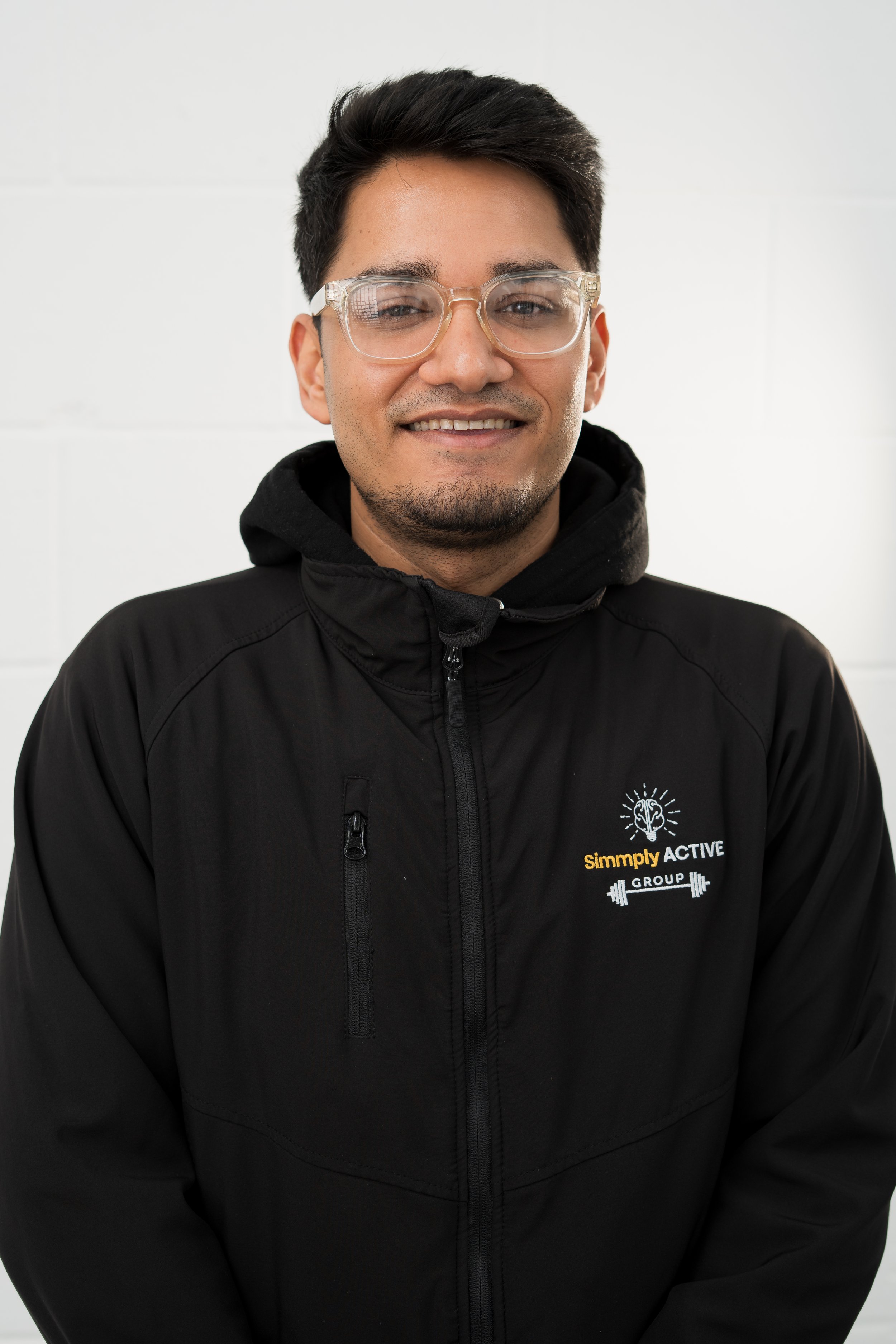 Man wearing glasses, a black jacket with 'Simply Active' logo, standing in front of a branded backdrop.