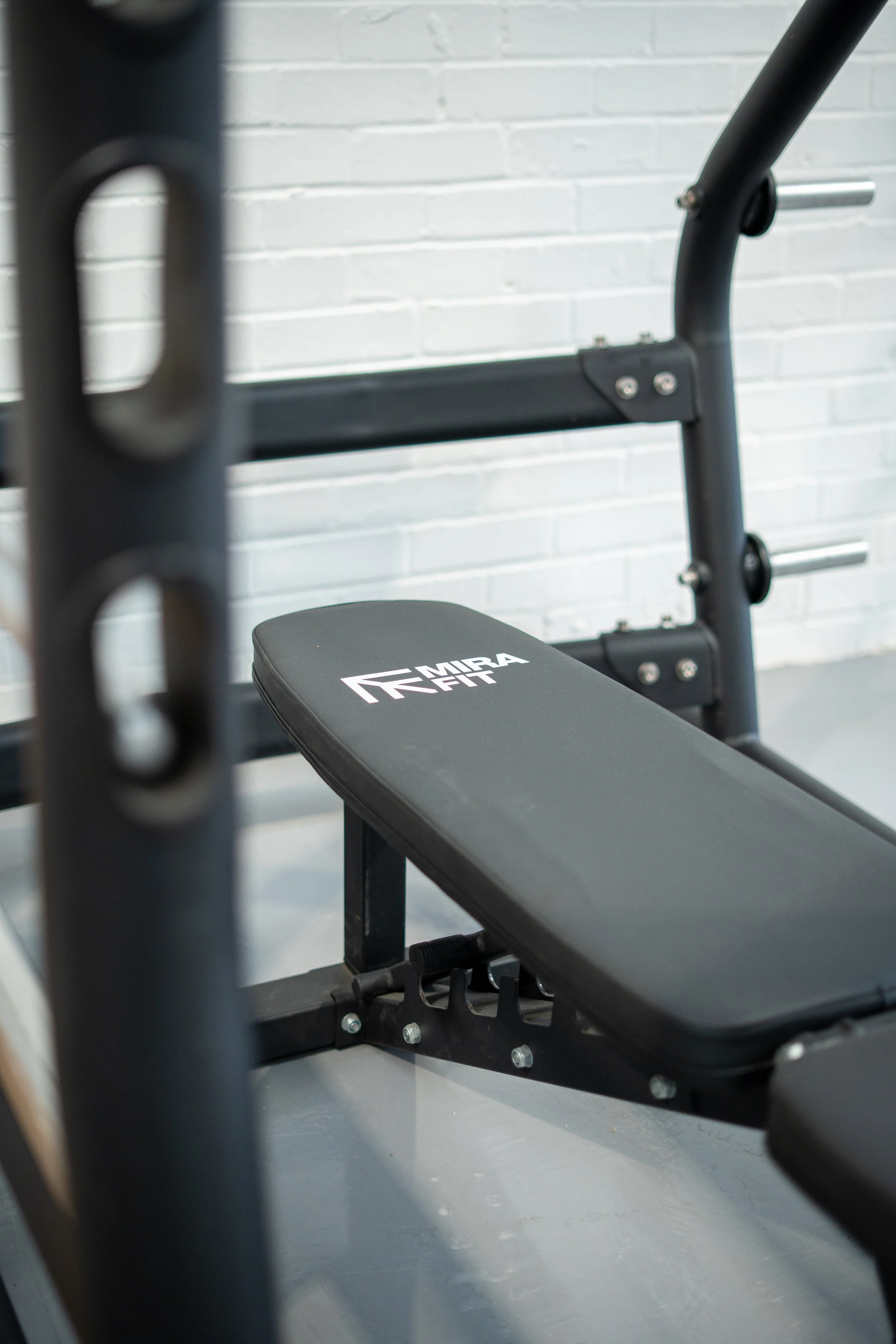 Close-up of a black weight bench with the brand name 'MIRA FIT' printed on the padding, positioned beside a white brick wall.