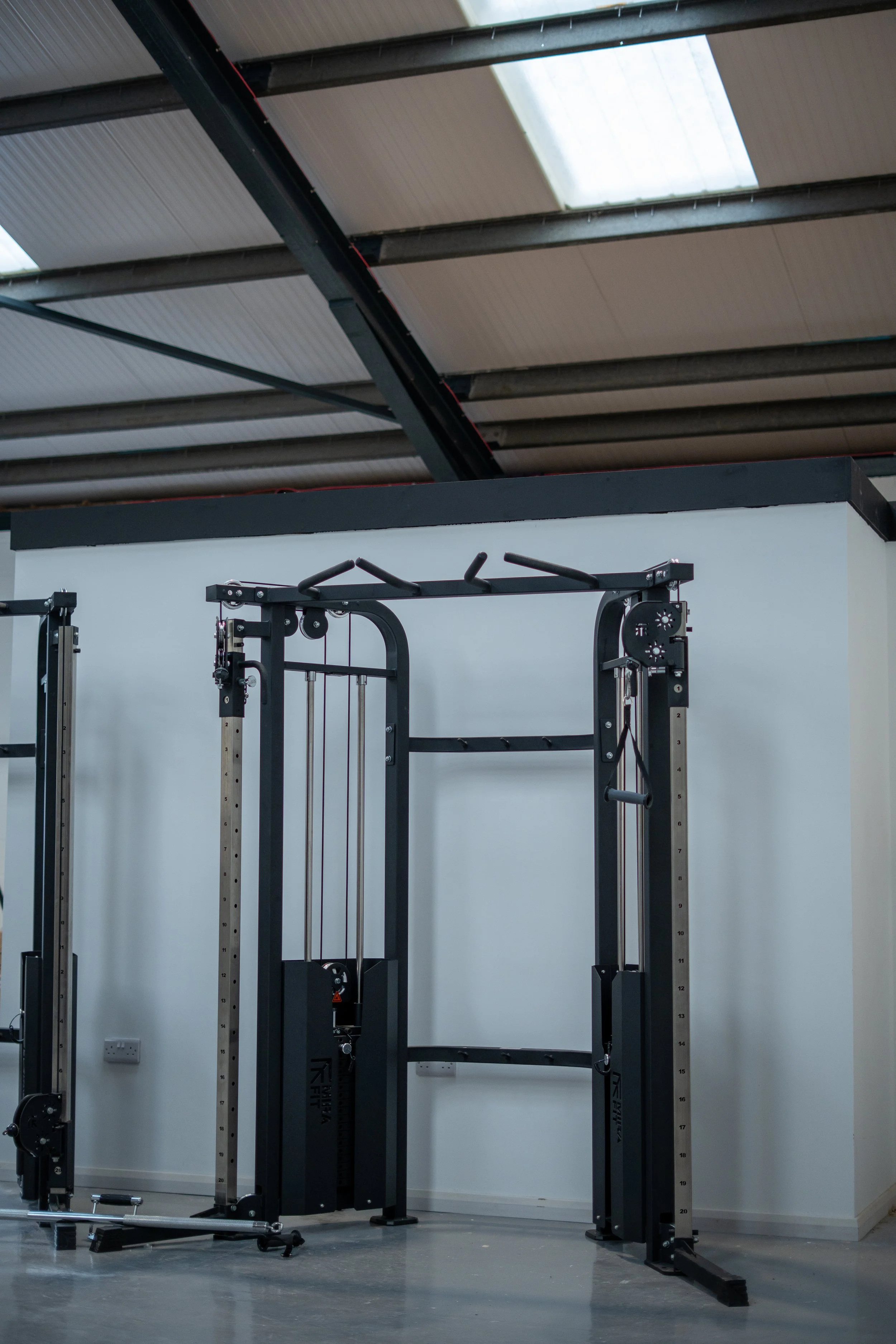 A gym machine with weight stack and cables in a fitness studio, with a white wall and a ceiling with skylights.