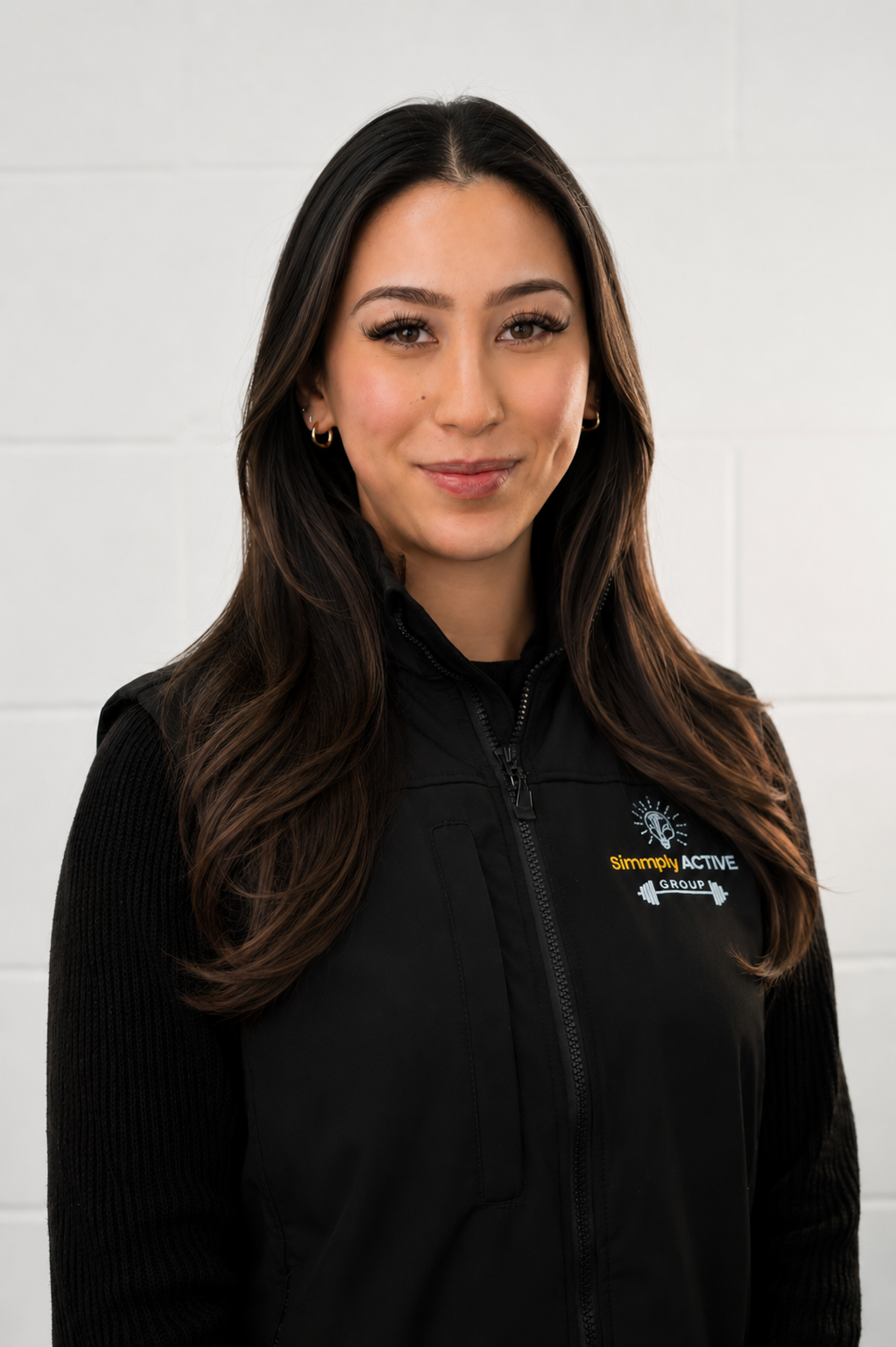 Portrait of a young woman with long dark hair, wearing a black zip-up jacket with a logo that says "Simply Active Group," standing against a white brick wall.