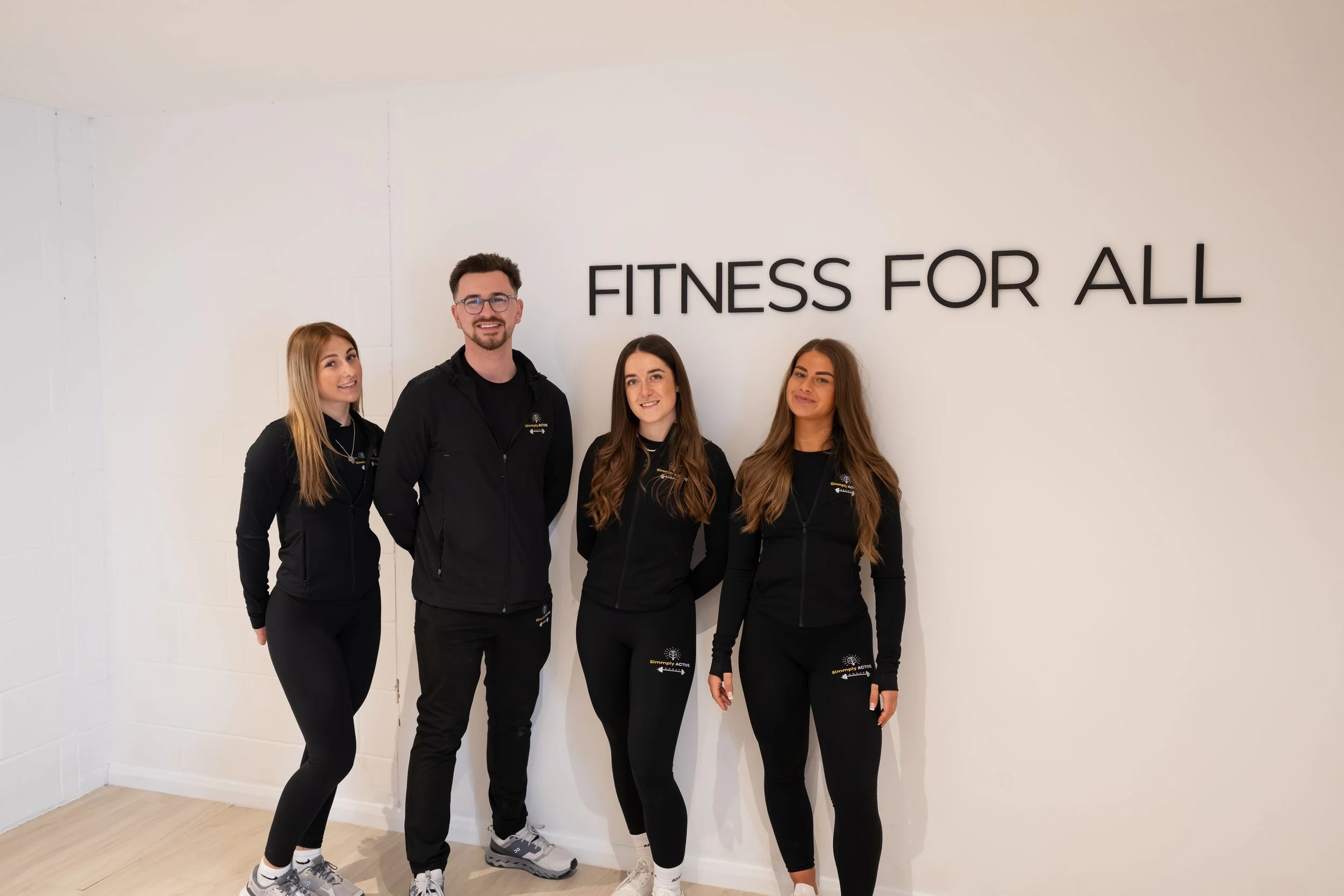 Group of four people standing indoors in front of a wall with the sign "Fitness for All". They are dressed in black activewear, smiling, and looking at the camera.