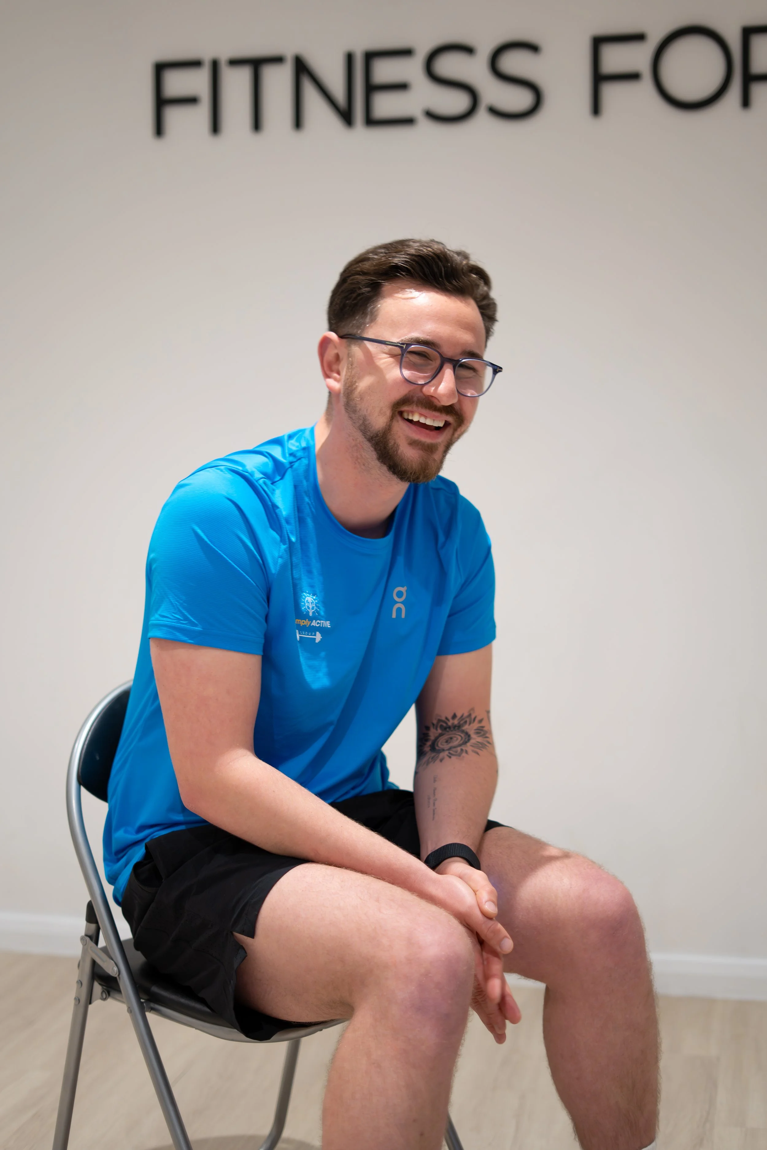 A young man with glasses and a beard, wearing a blue athletic shirt and black shorts, sitting on a chair and smiling in a room with a sign that reads 'Fitness for' in the background.