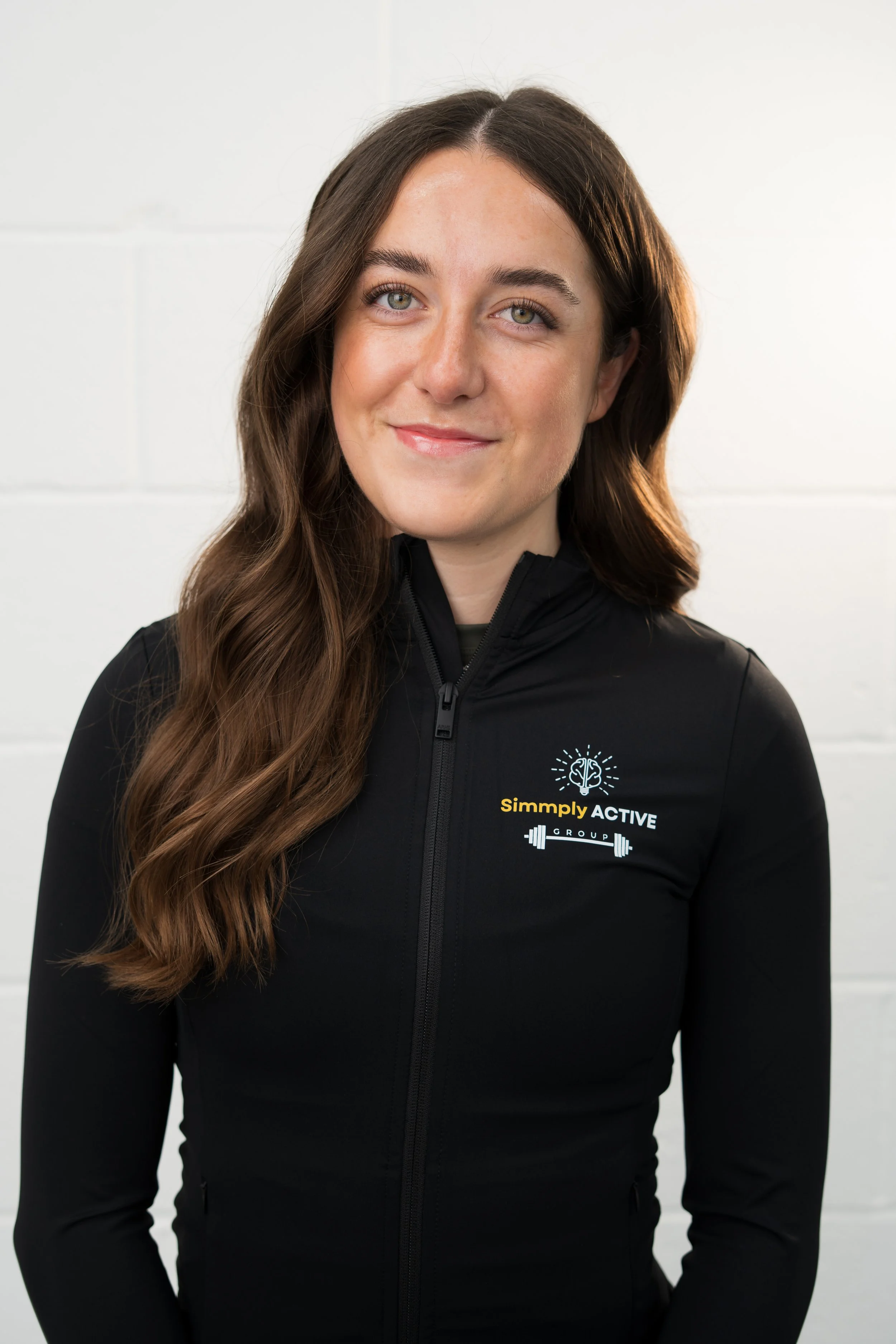 A woman with long brown hair smiling, wearing a black t-shirt and black leggings, standing in front of a white background with the 'Simply ACTIVE Group' logo.