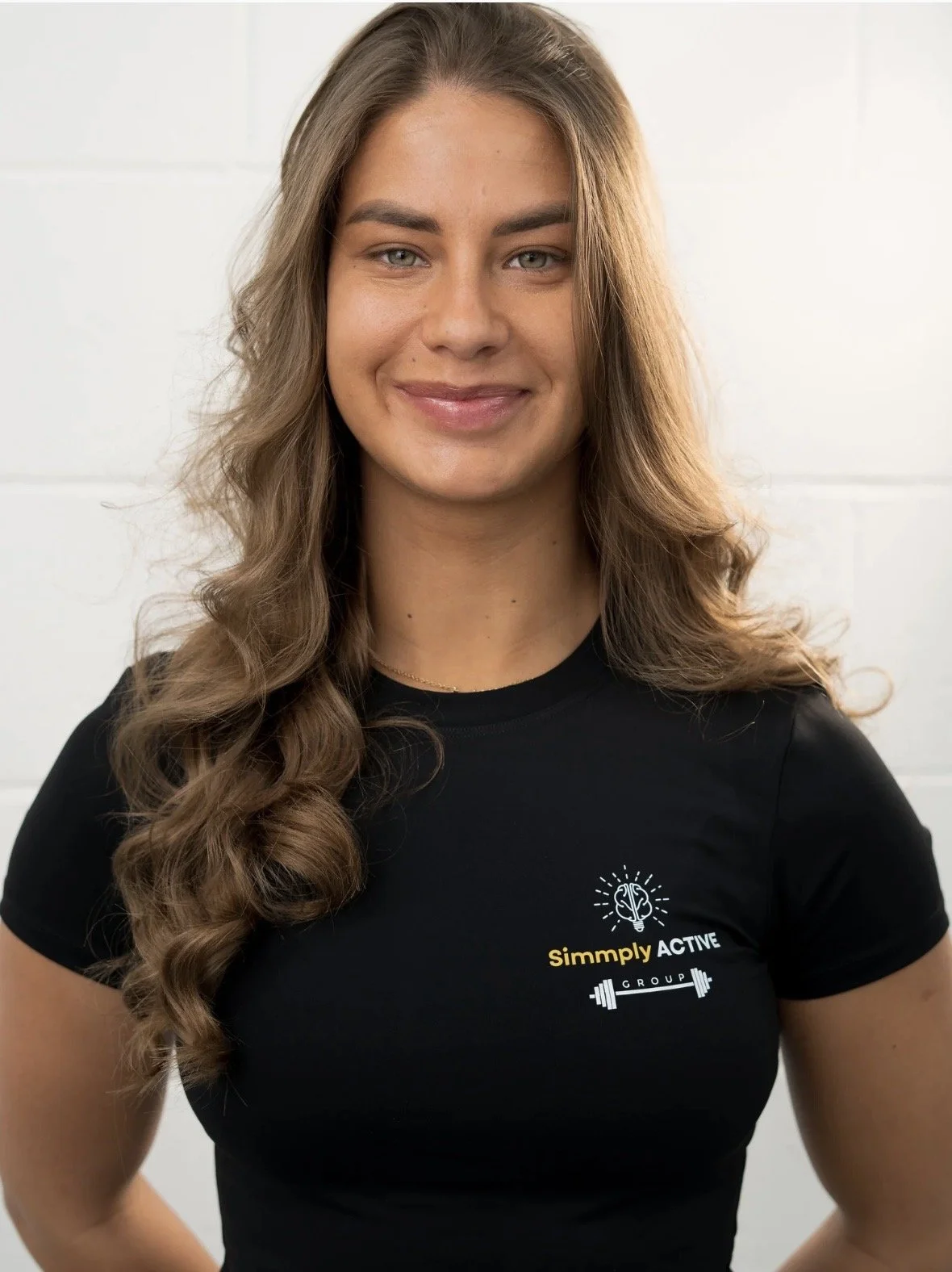 Young woman with long wavy hair wearing a black t-shirt with 'Simply ACTIVE' logo, standing against a white wall.