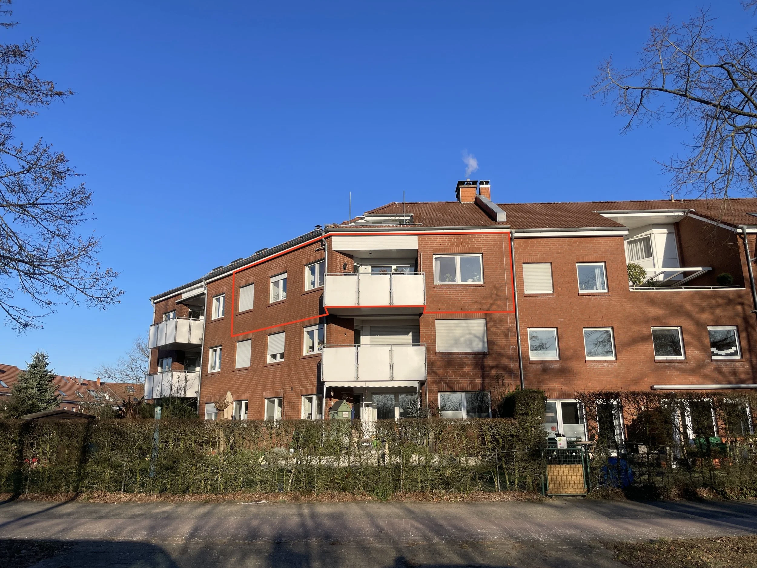 Mehrstöckiges rotes Backsteingebäude mit Balkonen, unter klarem blauen Himmel, im Vordergrund ein grüner Gartenzaun.