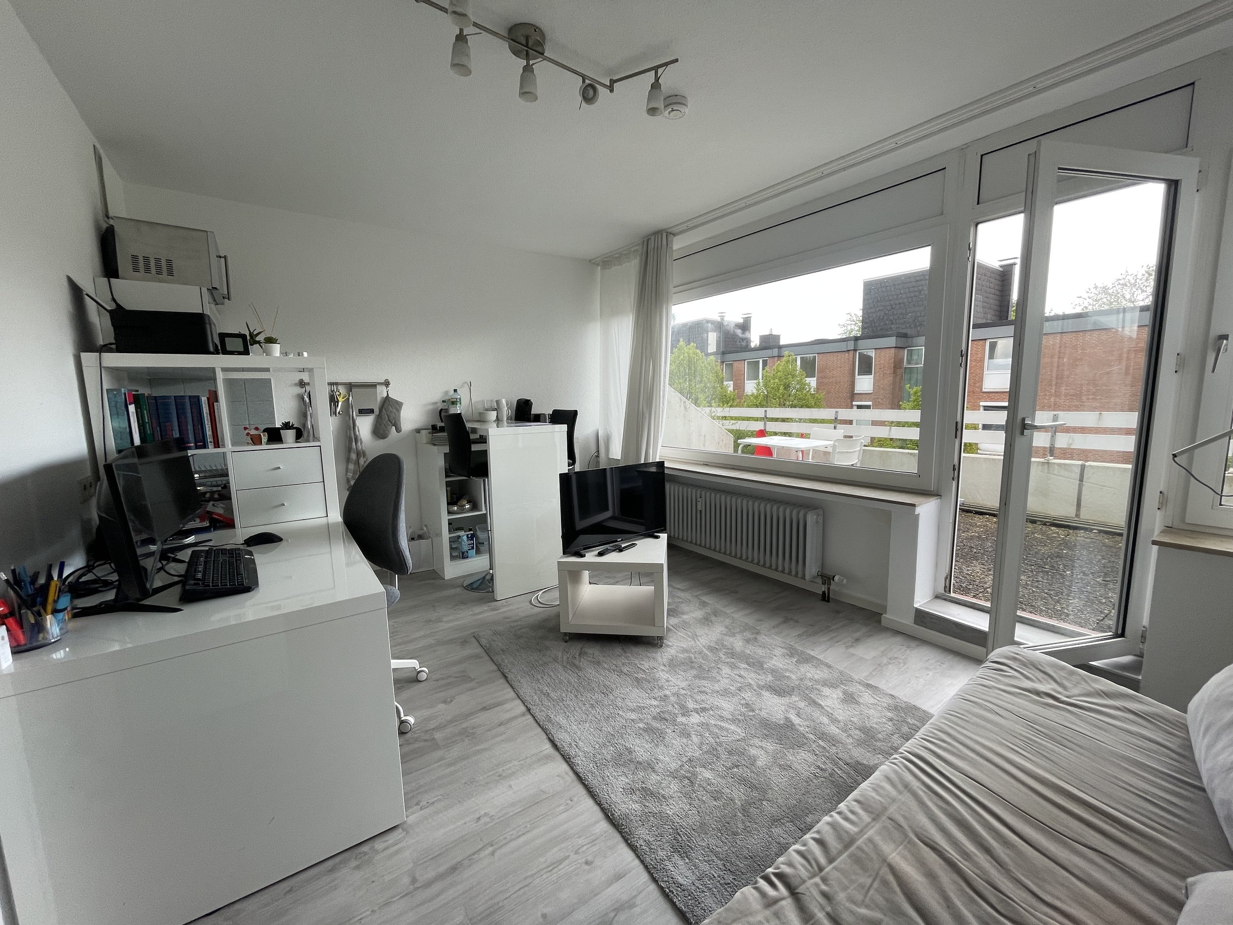 Ein Wohnzimmer mit Schreibtisch, Computer, Fernseher, Sofa, großen Fenstern und einer Balkonterrasse.