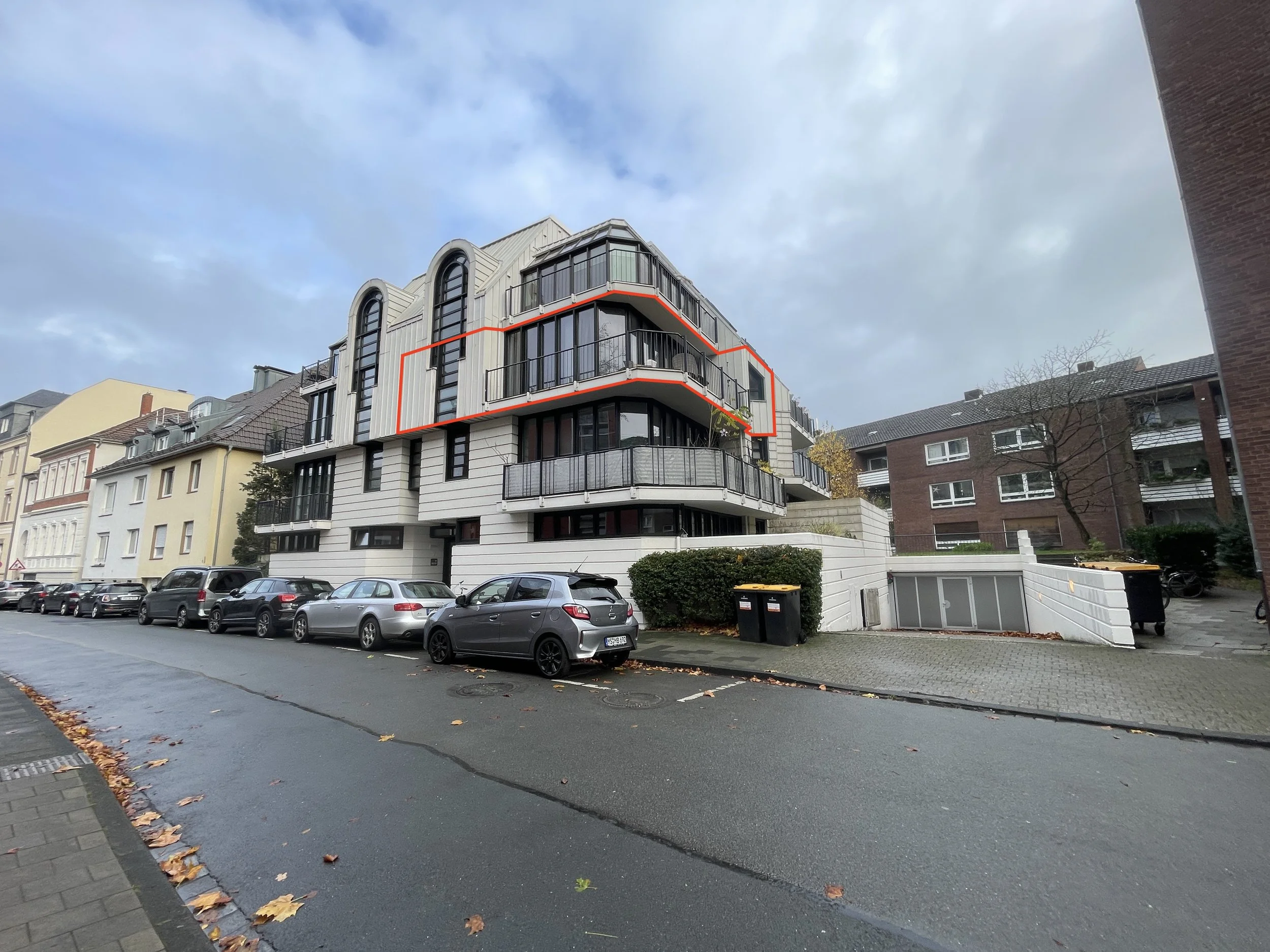 Mehrstöckiges modernes Wohngebäude mit großen Fenstern und Balkonen, Parkplätze im Vordergrund, bewölkter Himmel.