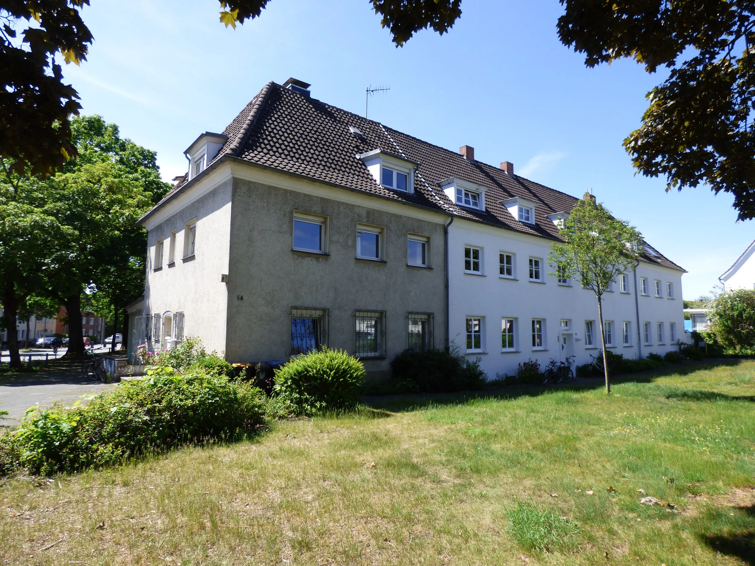 Mehrfamilienhaus mit weißen und grauen Fassaden, mehreren Fenstern, einem roten Ziegeldach und Bäumen im Garten bei sonnigem Himmel.