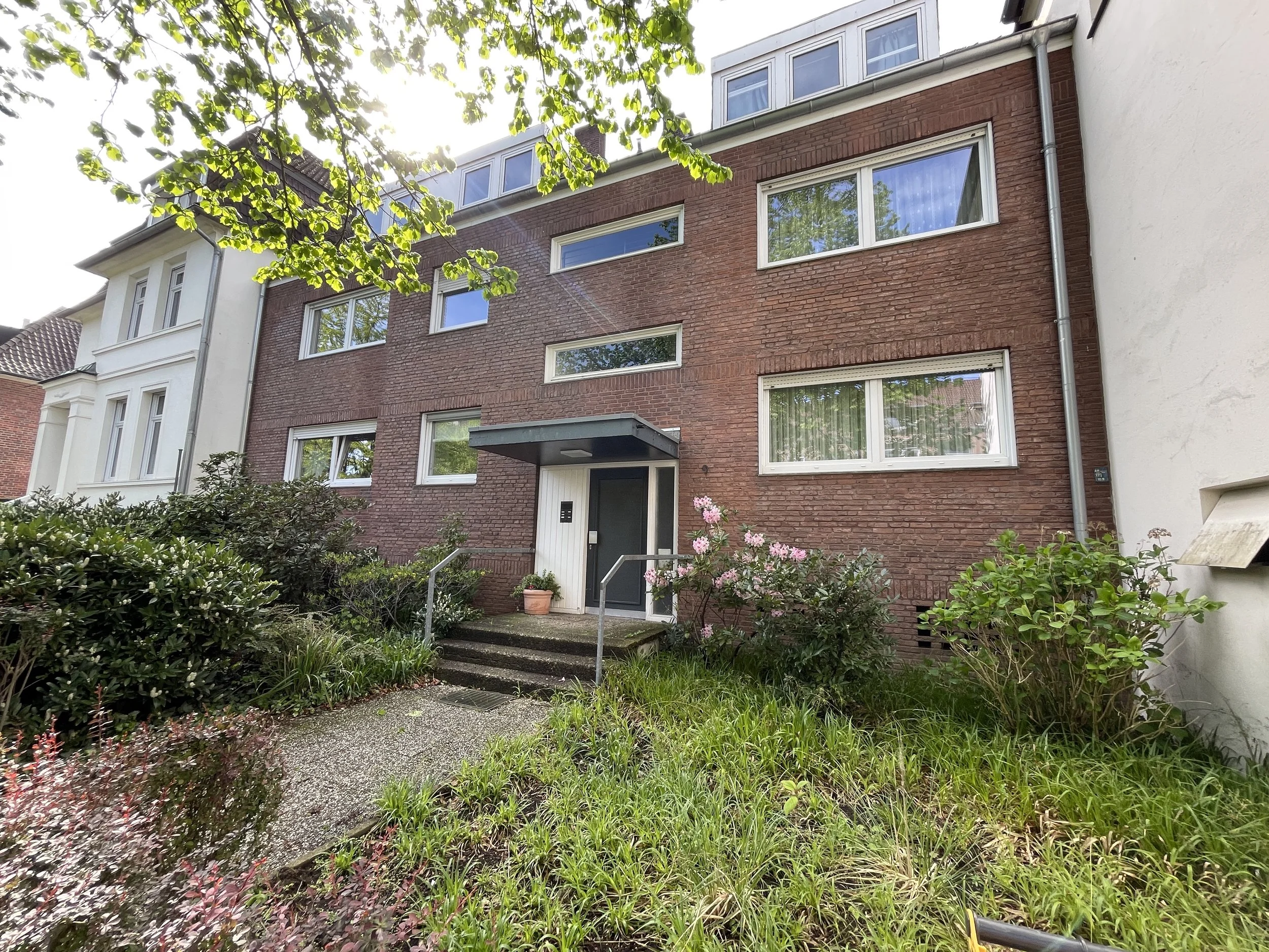 Mehrstöckiges Wohnhaus mit roten Ziegelsteinen, weißen Fenstern, Eingang mit Treppe, Garten mit Pflanzen und Bäumen.