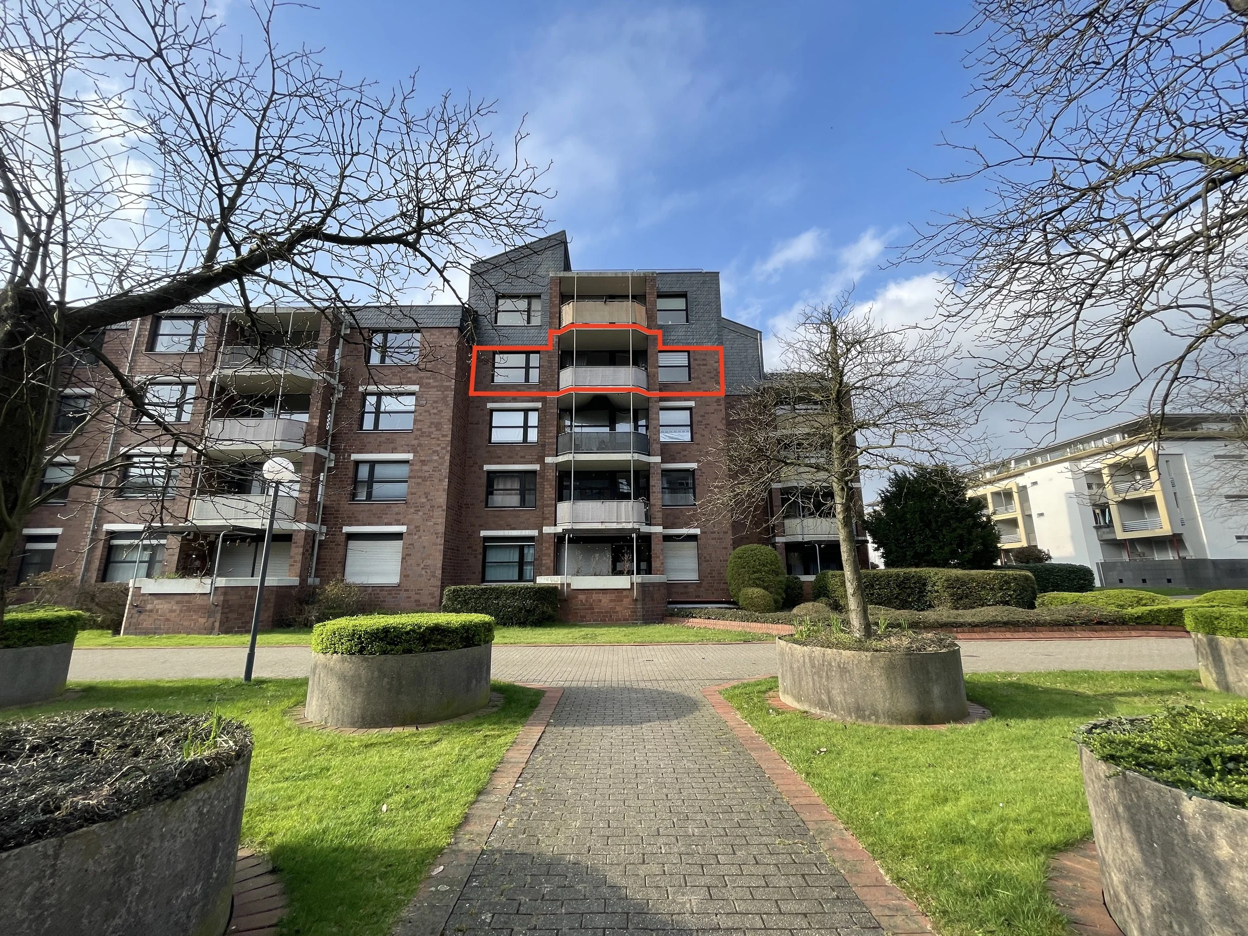 Mehrfamilienhaus mit Balkon und Bäumen im Vorgarten, blauer Himmel mit Wolken.