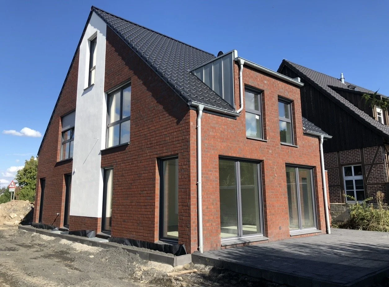 Neues modernisiertes Haus mit roten Ziegelwänden, großen Fenstern und einem dunklen geneigten Dach, im Hintergrund blauer Himmel.
